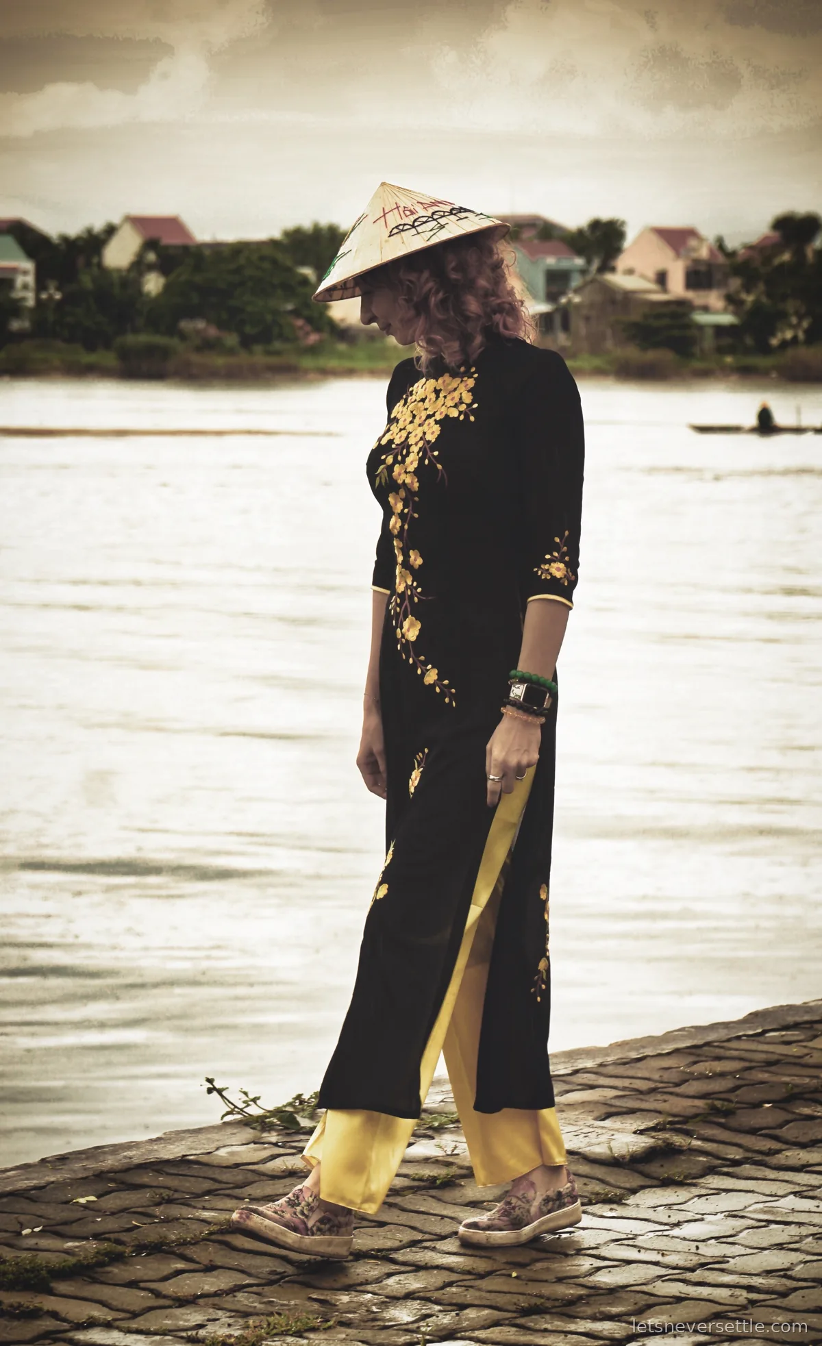 Inga in a black ao dai with yellow embroidery and matching yellow trousers, wearing a conical hat printed Hoi An, walking along a cobbled riverbank with houses on the far shore