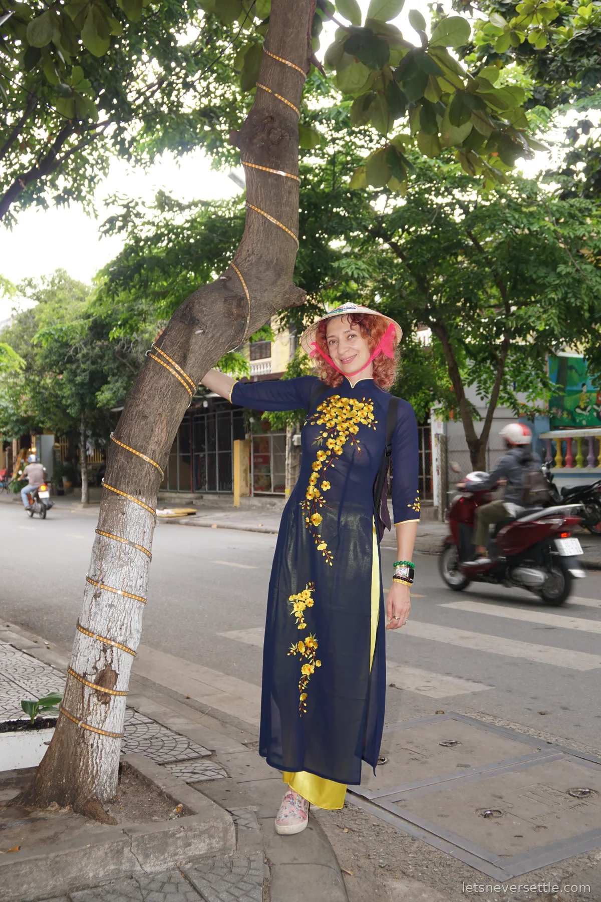 Inga in a navy ao dai embroidered with yellow blossoms and a pink-trimmed conical hat, leaning against a tree trunk wrapped in gold ribbon on a quiet street