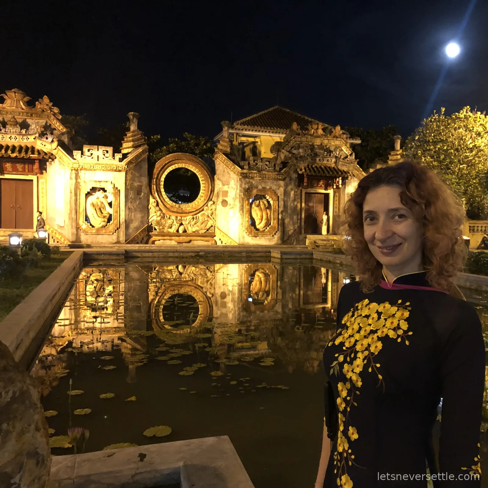 Inga in a dark ao dai embroidered with yellow blossoms beside a lily pond that reflects a golden illuminated temple gate at night, a full moon above