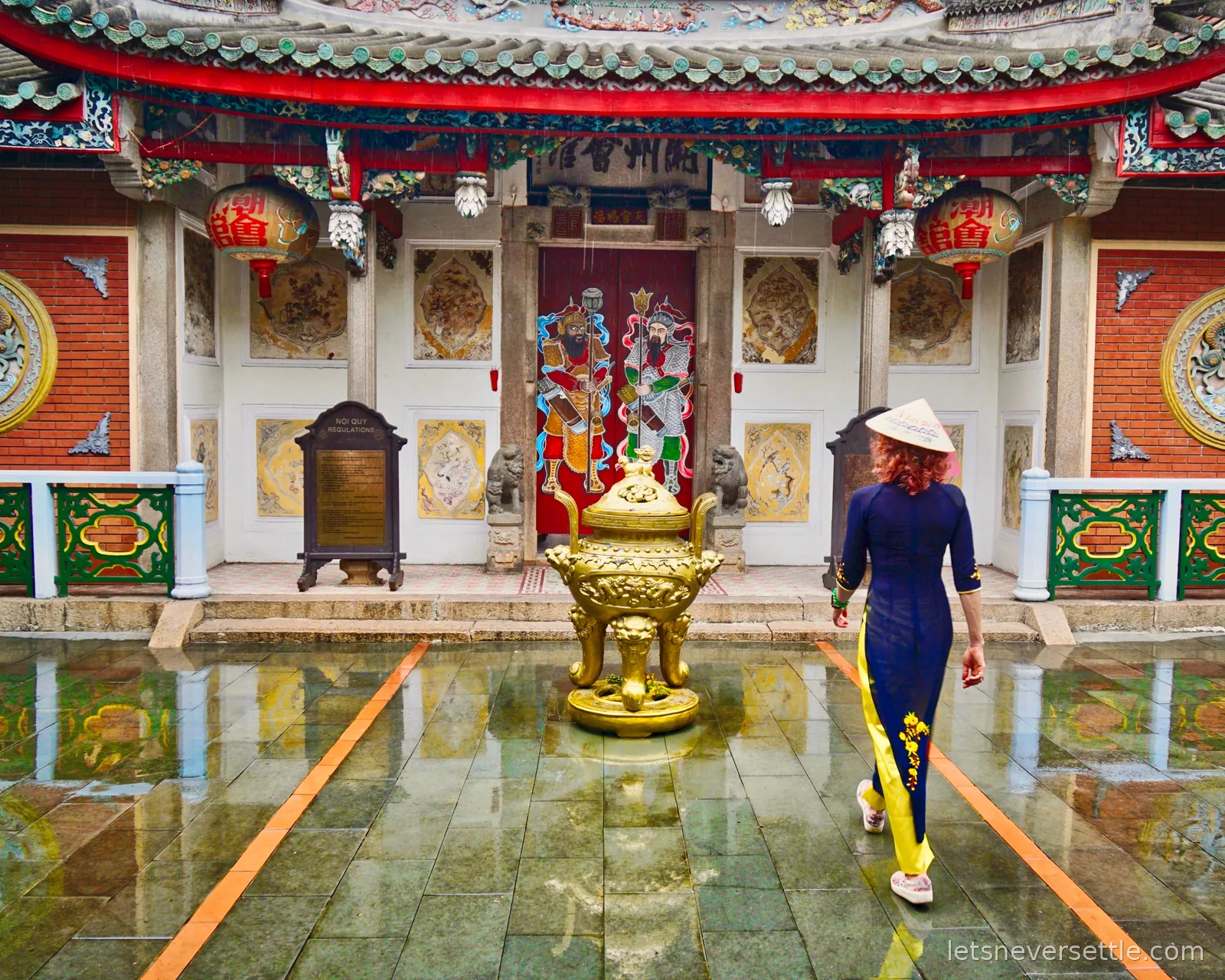 Inga in a navy and yellow ao dai and conical hat walking across a rain-soaked stone courtyard towards a Chinese assembly hall with a golden incense urn, painted door guardians and red lanterns