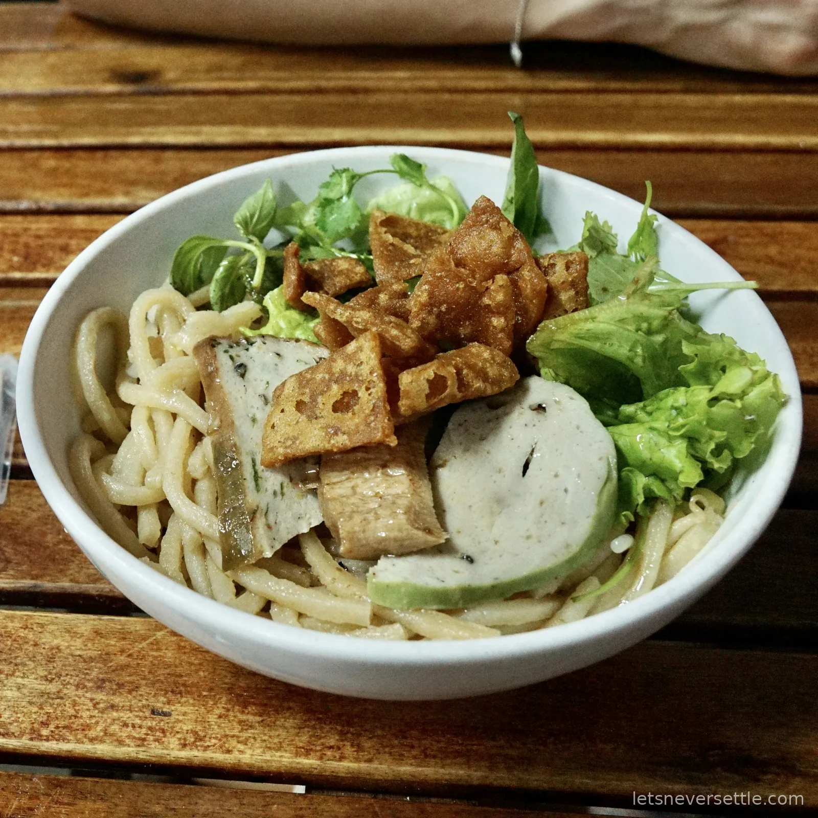 A white bowl of cao lau: thick pale noodles topped with sliced mock meat, crisp golden croutons and fresh green herbs and lettuce, on a slatted wooden table