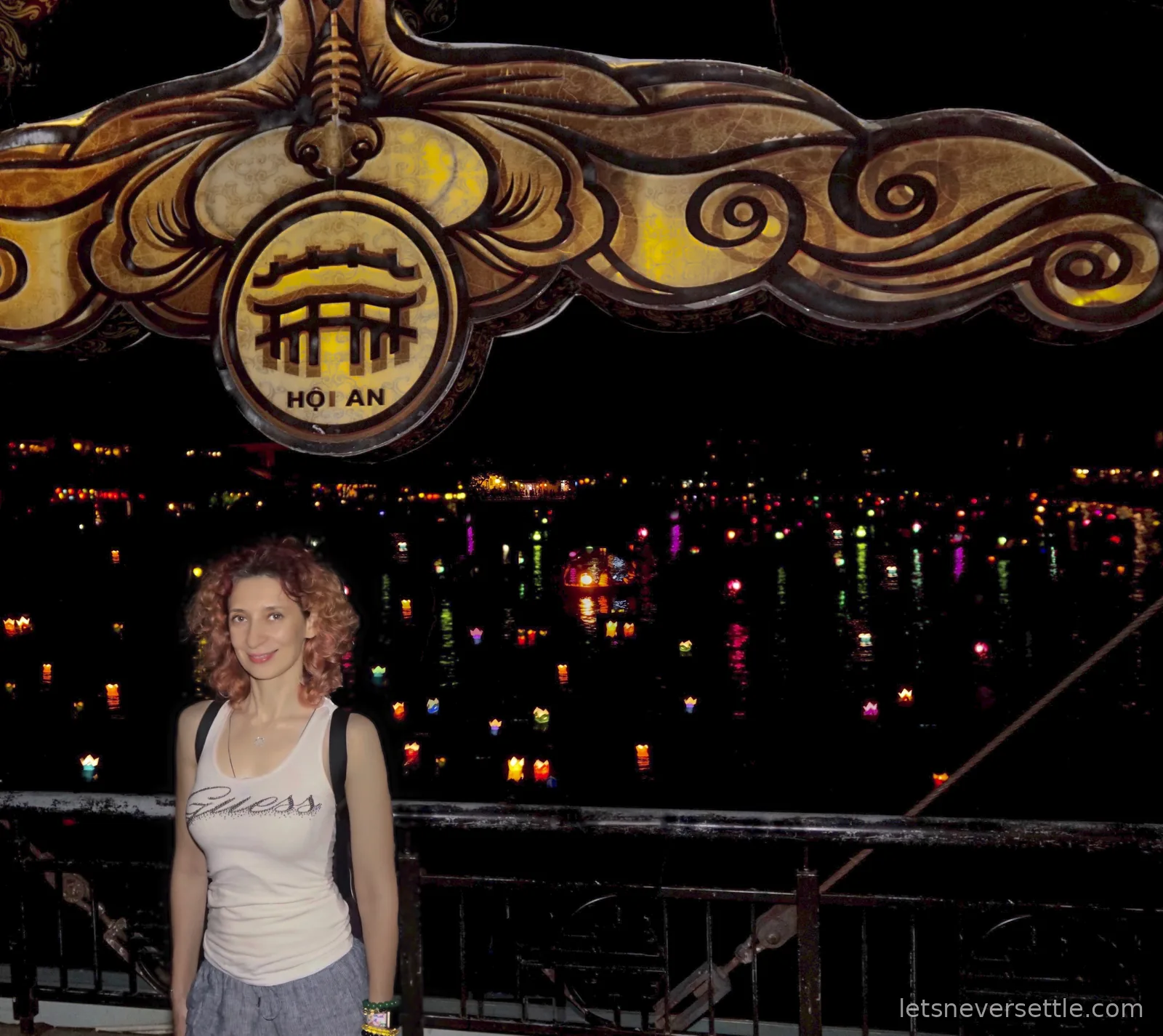 Inga standing at a railing beneath a large carved gold sign reading Hoi An, with hundreds of small coloured floating lanterns scattered across the dark river behind her