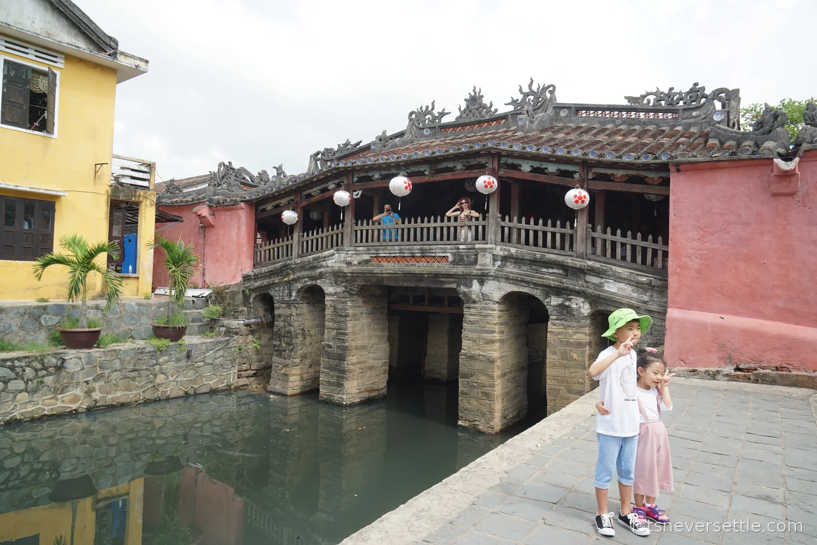 Side view of the Japanese covered bridge showing its stone arches over the canal, white paper lanterns hanging inside, and two children posing on the paving in front