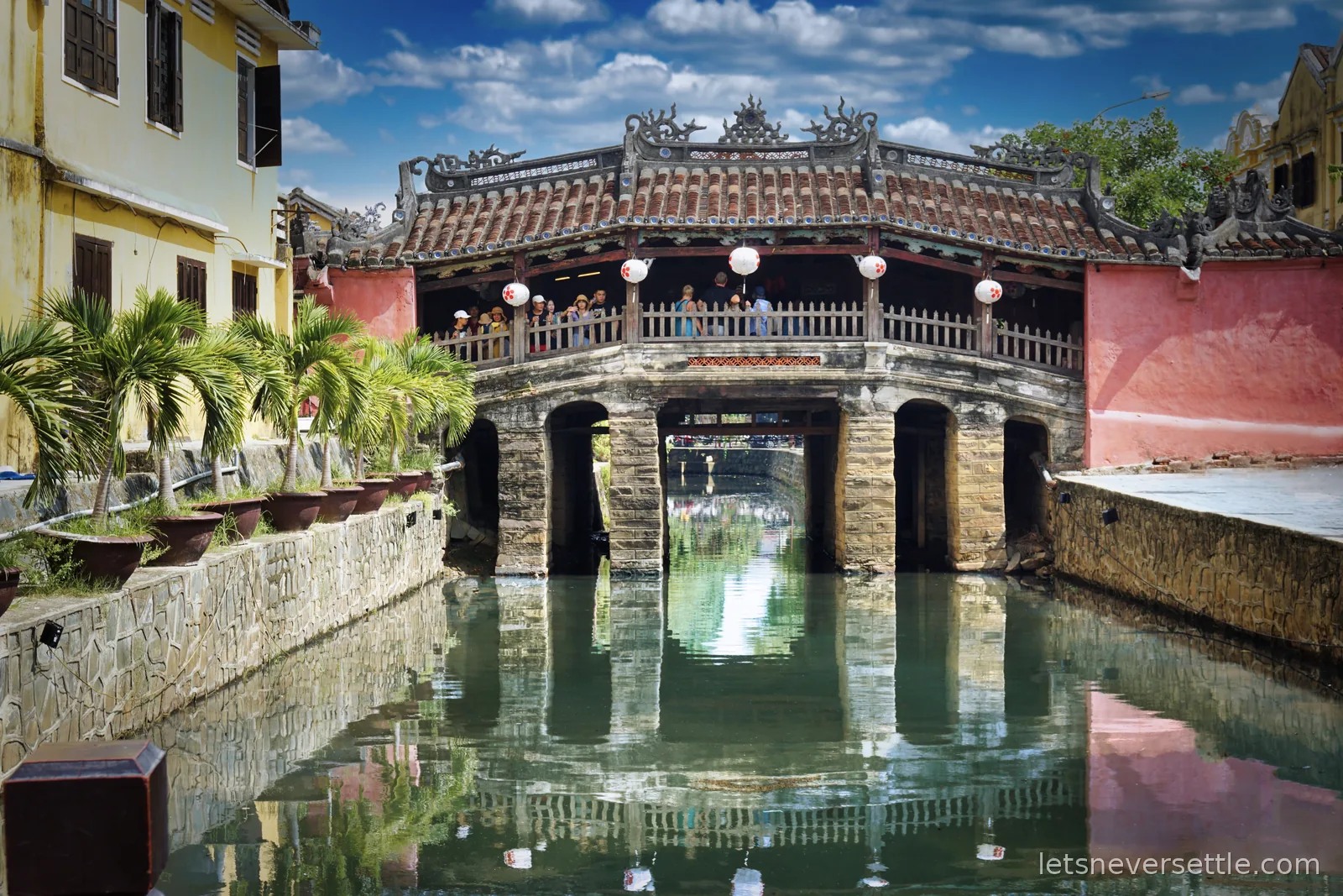 The Japanese covered bridge in Hoi An seen head-on across a green canal, its tiled roof and carved ridge reflected in the water, with a yellow building on the left and a pink wall on the right