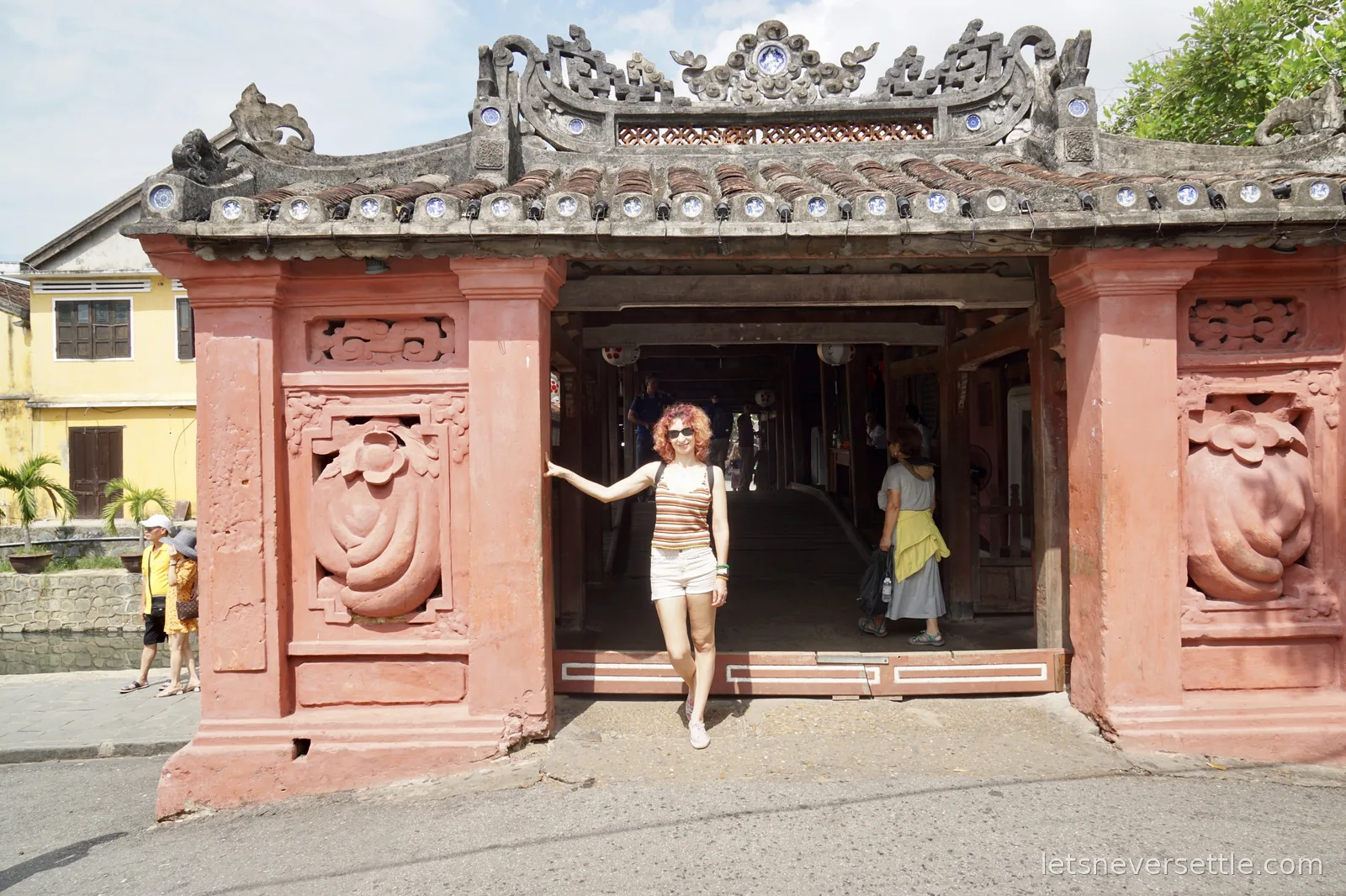 Inga standing in the pink entrance archway of the Japanese covered bridge, with carved stone panels either side and the dark wooden interior behind her