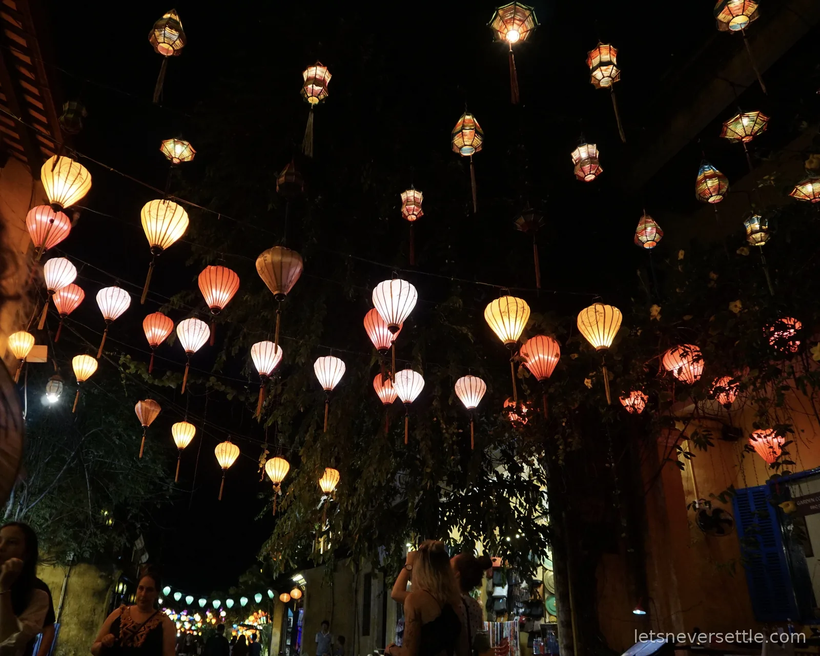Dozens of teardrop-shaped silk lanterns in pink, cream and yellow hanging on wires above a night street in Hoi An, lit from within against a black sky