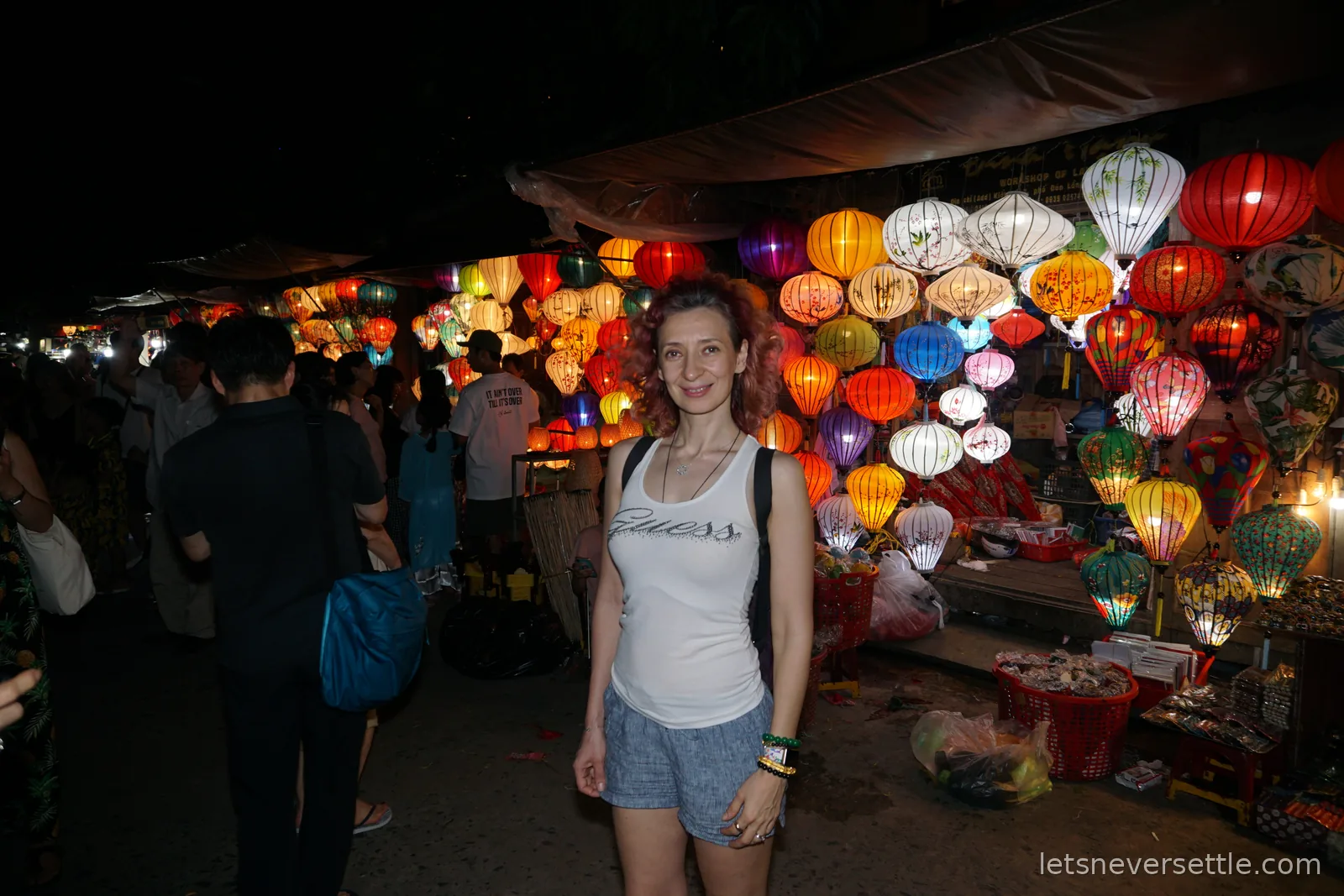 Inga standing in front of a night market stall packed with hundreds of glowing silk lanterns in red, orange, green and white, with shoppers passing on the left