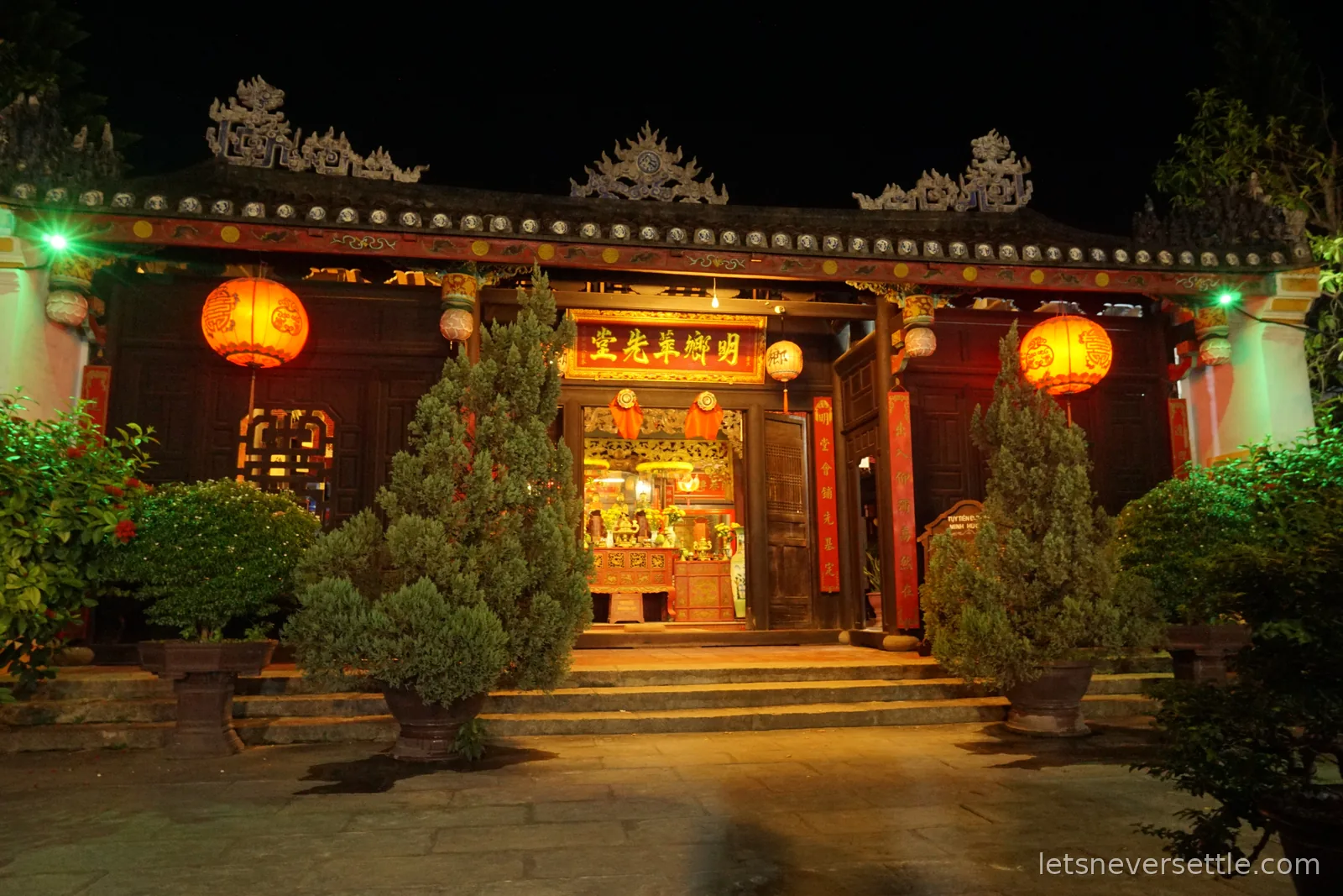 The lit facade of a Chinese communal house in Hoi An at night, with red lanterns either side of the entrance, gilded characters above the door and clipped conifers in pots on the steps