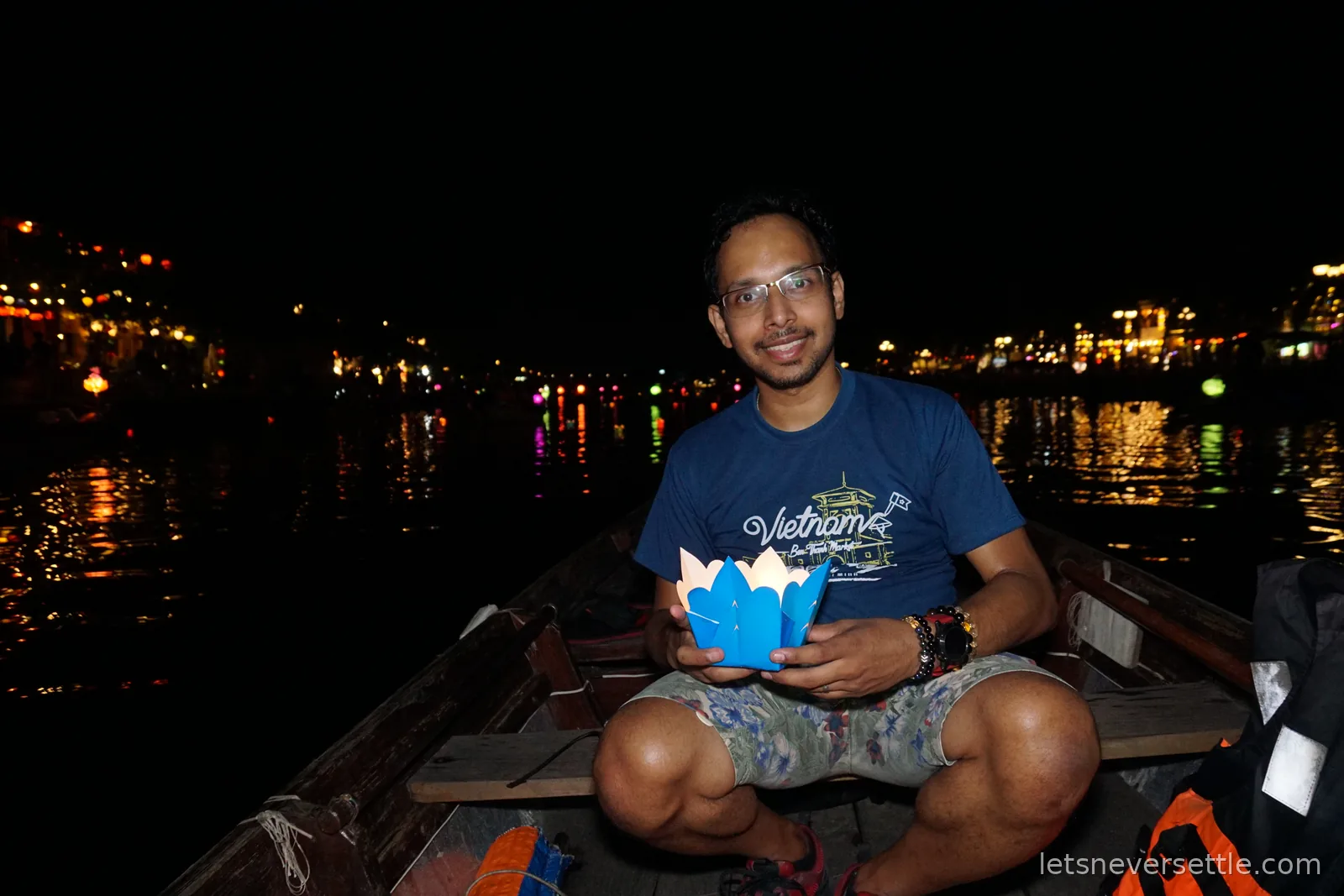 Dileep sitting cross-legged in a small wooden rowing boat on the Thu Bon river at night, holding a blue paper lotus lantern, with the lit riverfront behind him