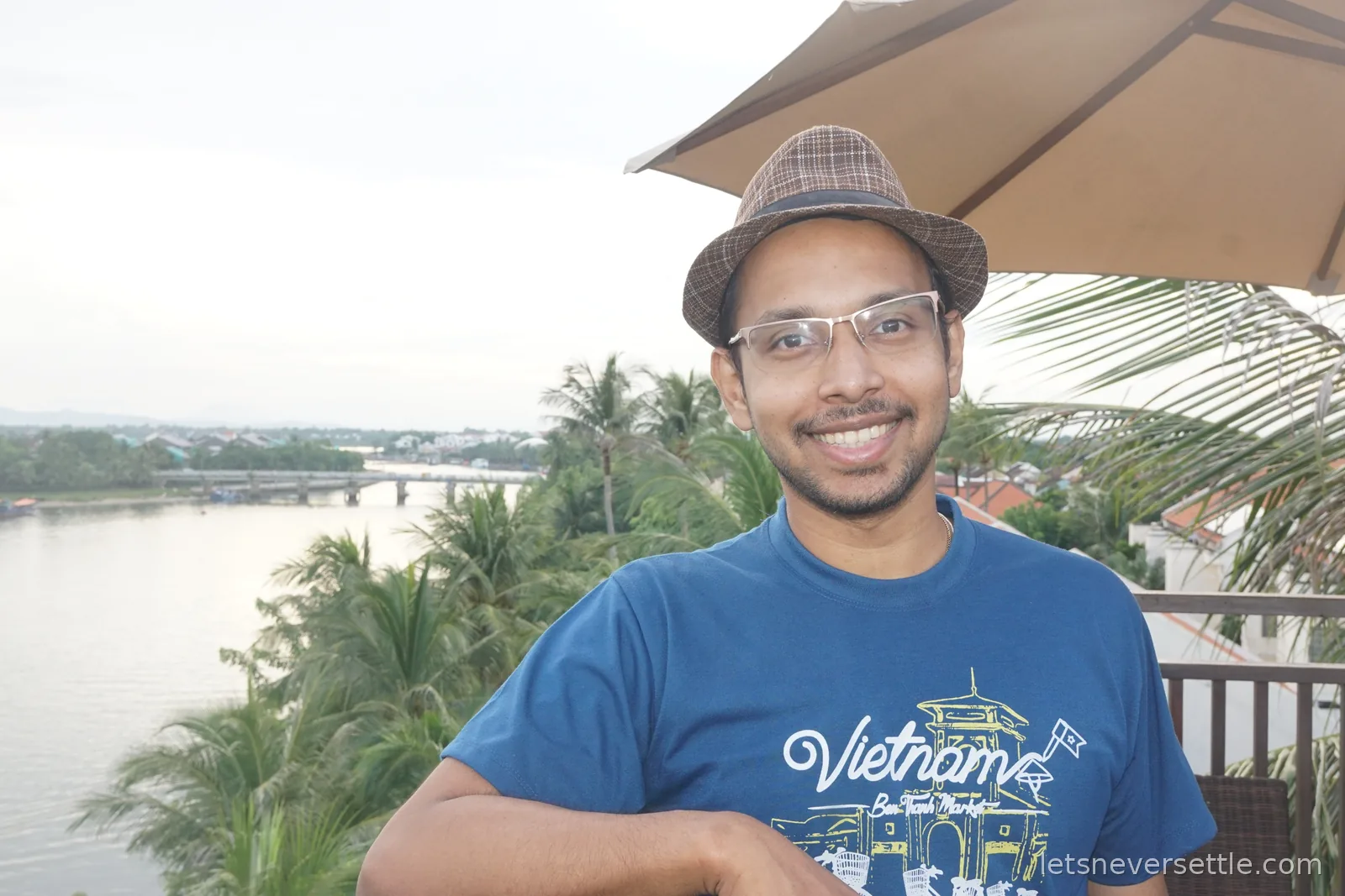 Dileep in a straw trilby and glasses smiling on a terrace with a parasol overhead, palm trees and the Thu Bon river with a low bridge stretching out behind him