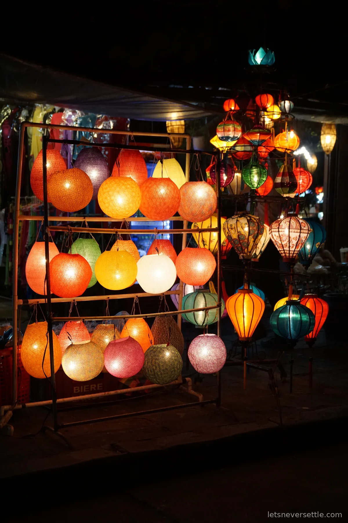 Round and teardrop silk lanterns in orange, pink, teal and white stacked on a metal frame in a dark alley, glowing against the shadows