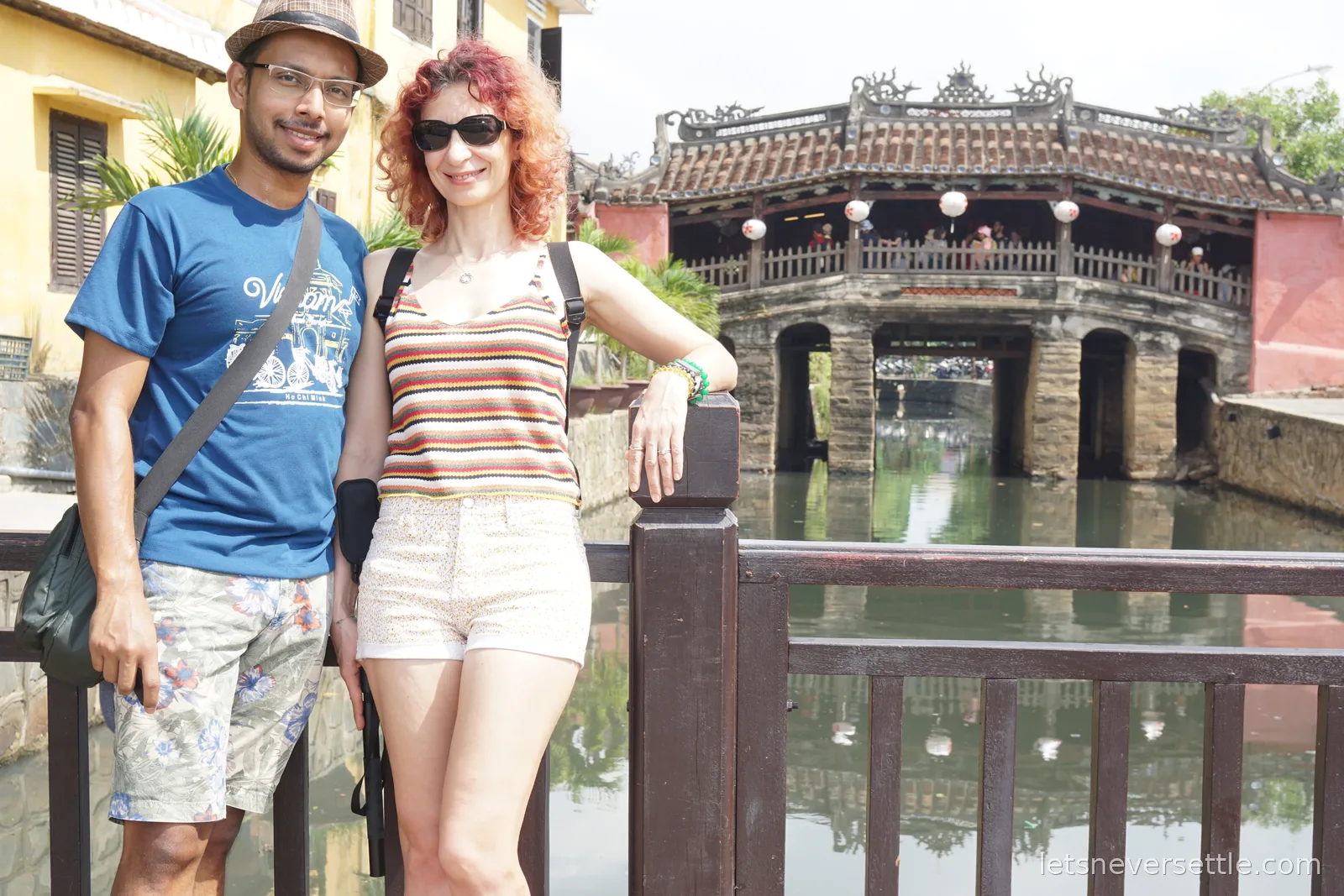 Inga and Dileep in sunglasses and a straw hat leaning on a dark wooden railing with the Japanese covered bridge and its stone arches behind them