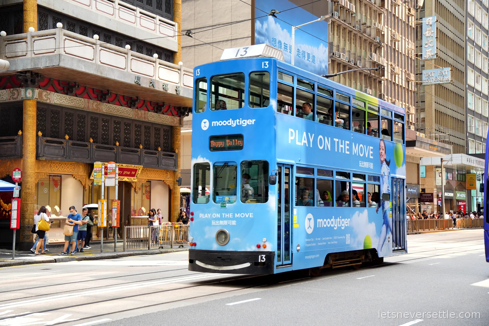 A blue double decker Hong Kong tram numbered 13 with Happy Valley on its destination blind, passing a gold-columned Chinese facade