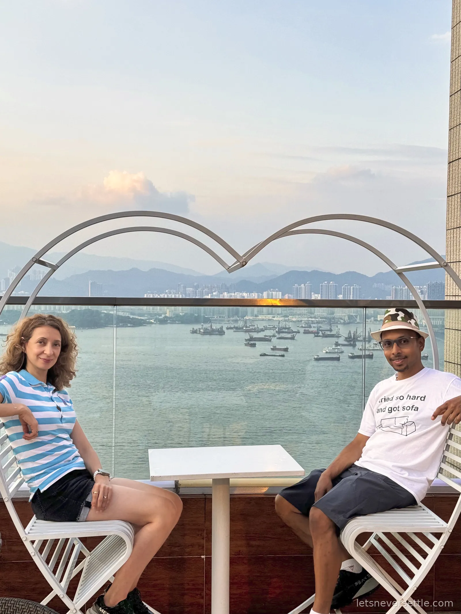 Inga and Dileep sitting at a white table on a hotel rooftop terrace under a heart-shaped metal arch, Hong Kong harbour and moored boats behind them