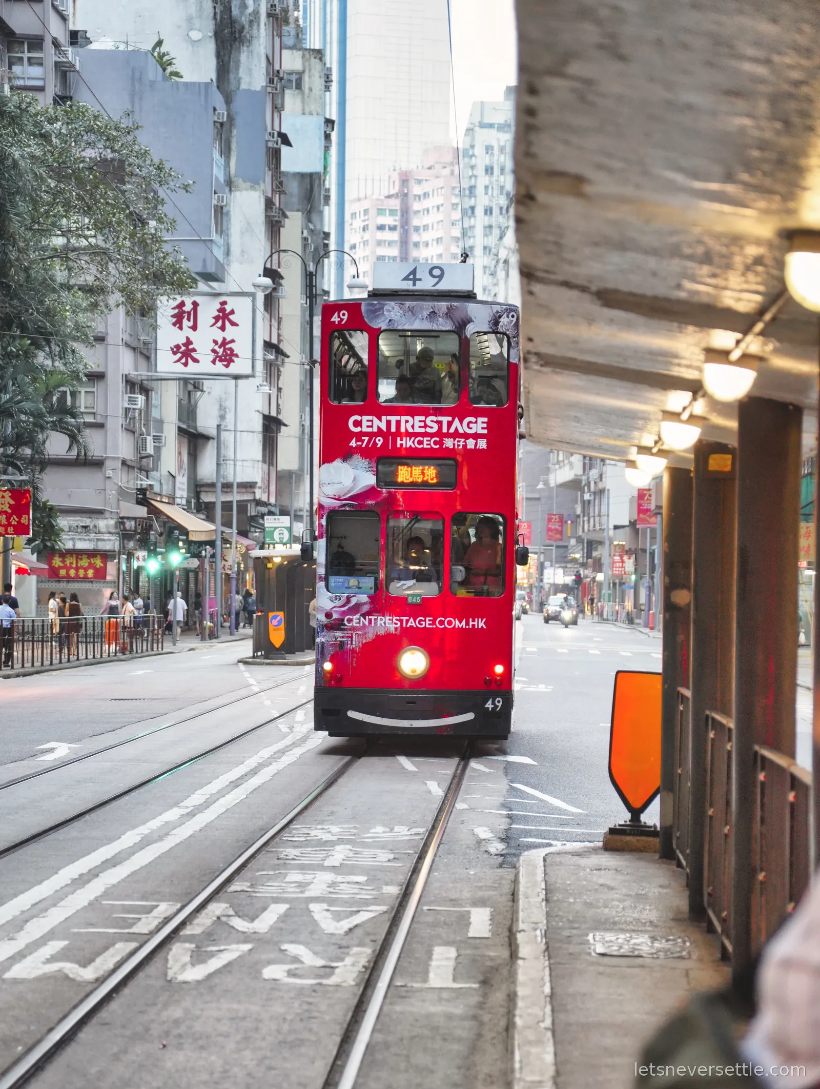A red double decker Hong Kong tram coming down the tracks between old shopfronts and Chinese signs, a tram lane marking painted on the road