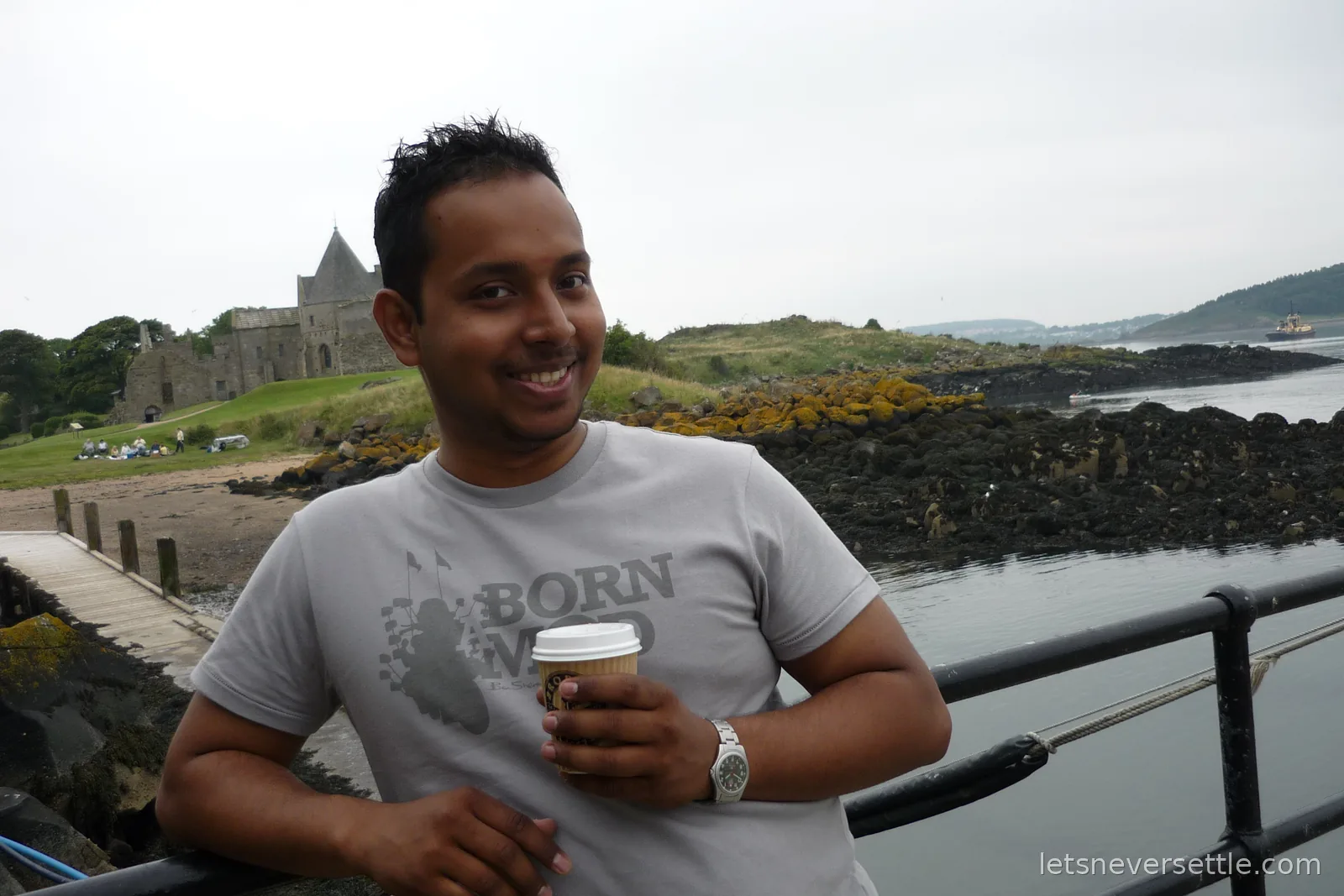 Dileep leaning on a railing by the Inchcolm jetty with a takeaway coffee, the stone tower of Inchcolm Abbey and the beach behind him
