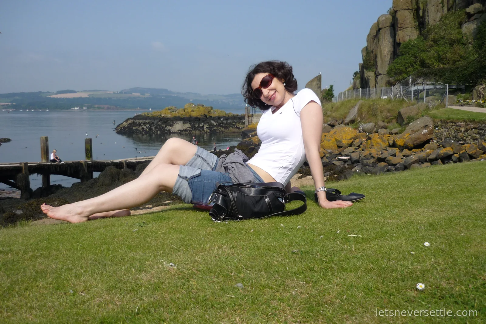 Inga sitting barefoot on the grass beside Inchcolm's rocky shore, with gulls over the water and the wooden jetty behind her