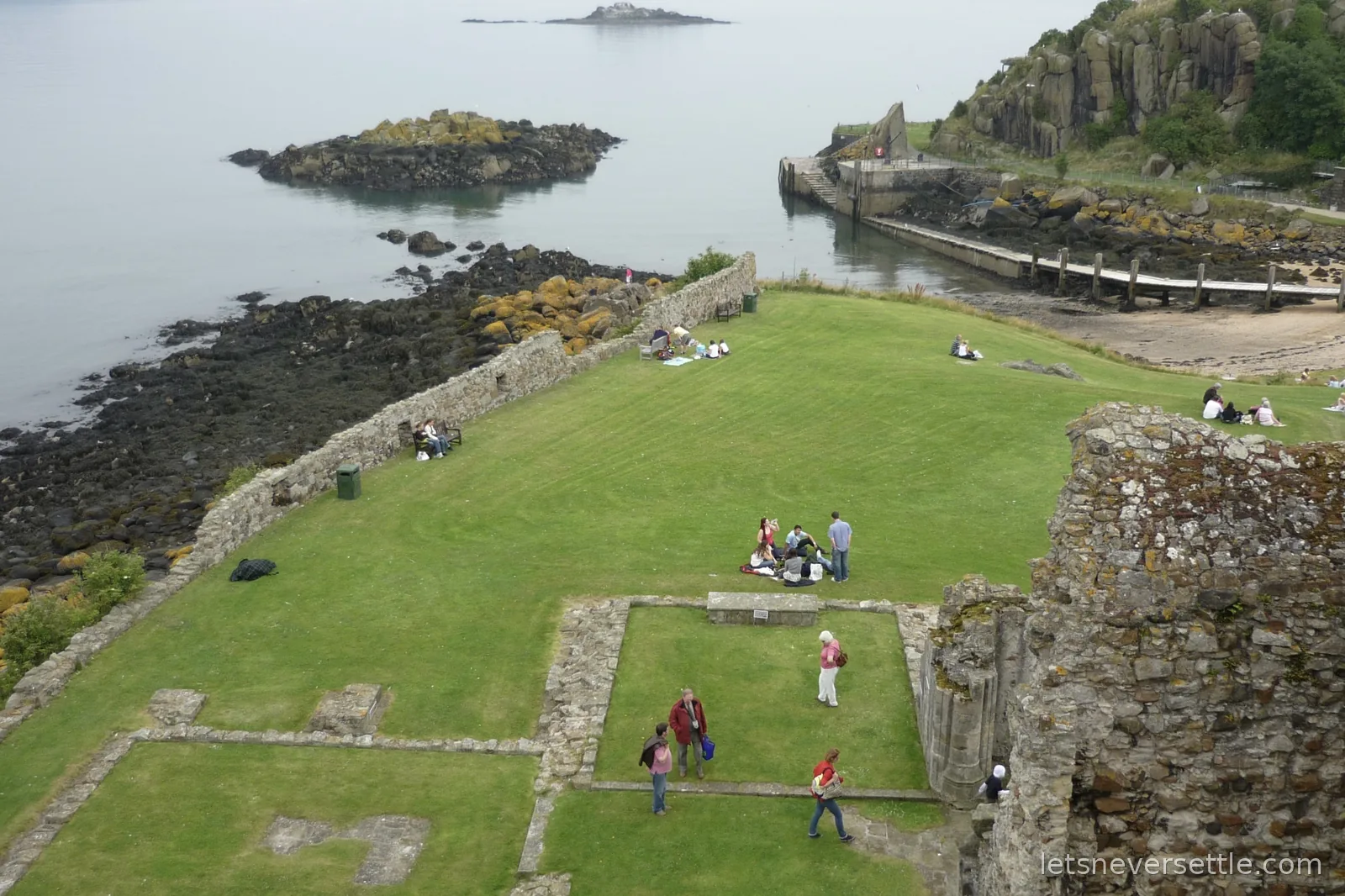 View from the top of Inchcolm Abbey tower over the ruined cloister walls, a green lawn dotted with picnicking visitors, and the island jetty on the rocky shore