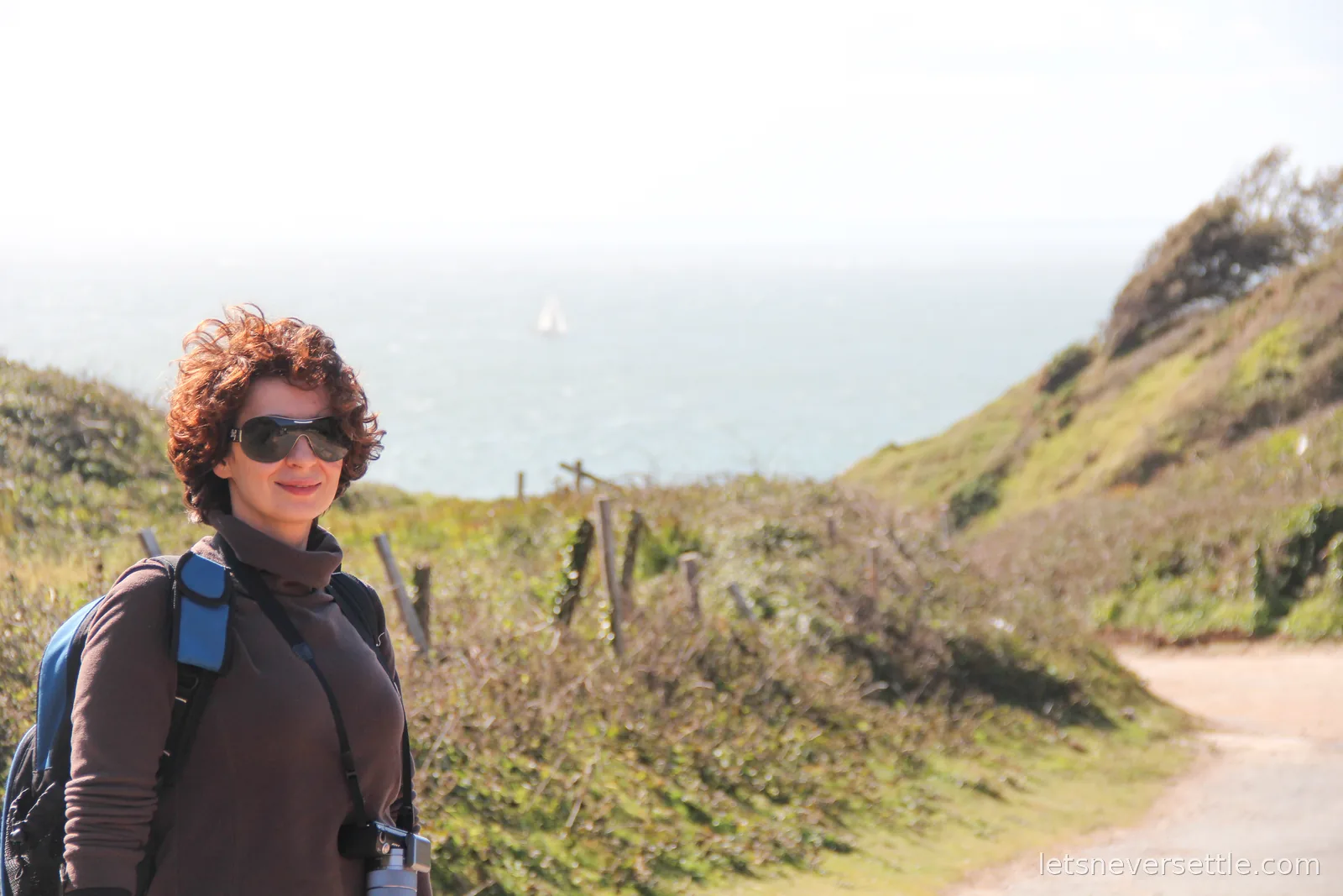 Inga on a clifftop path with hedgerows, a hazy sea and a distant white sail behind her