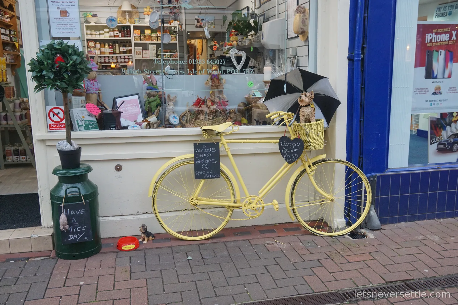 A bicycle painted entirely yellow, with a wicker basket and a chalkboard advertising cheese wedges, propped against a shopfront on Cowes High Street beside a green milk churn reading Have a Nice Day