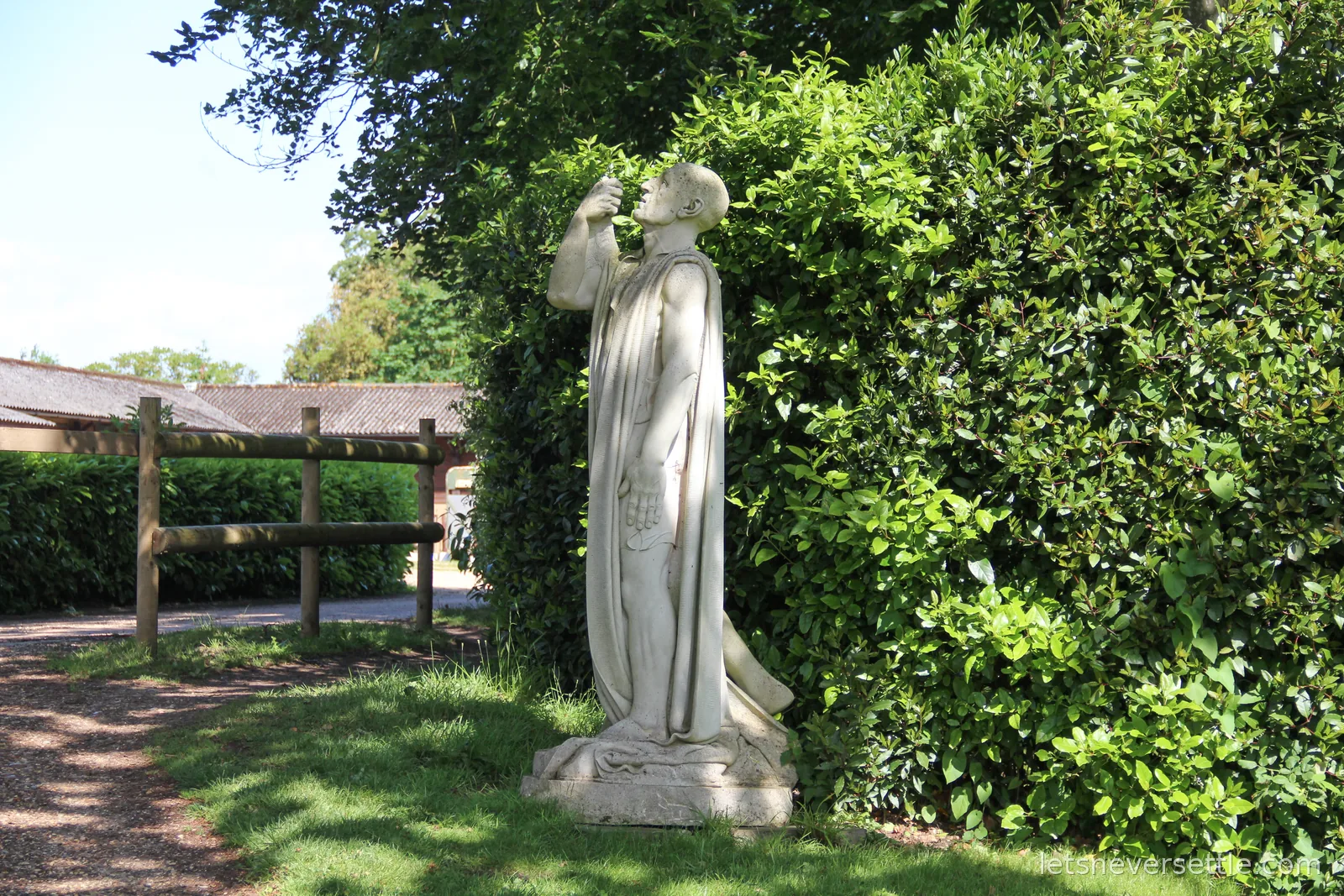 White stone statue of a robed monk drinking, set against a tall green hedge beside a fenced path
