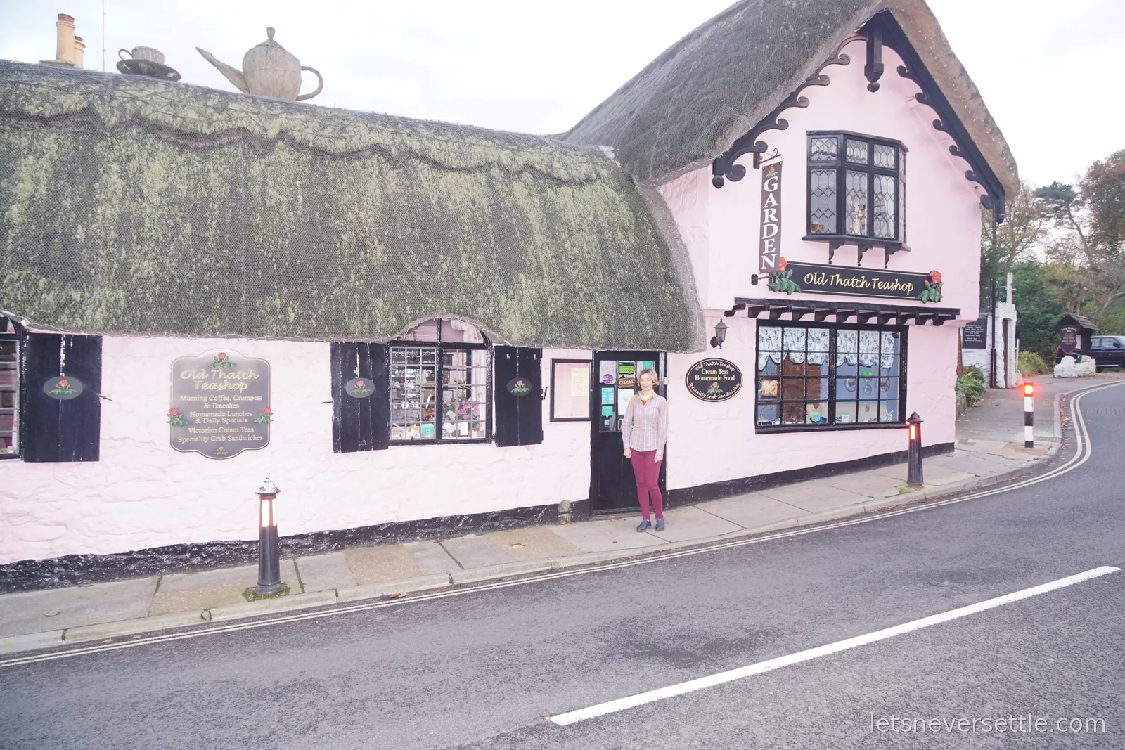 Inga standing in the doorway of the Old Thatch Teashop in Shanklin, a pink-painted building under a deep thatched roof with a giant thatched teapot and cup on the ridge