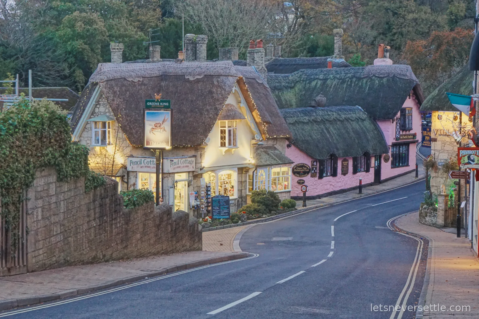 Pencil Cottage and the neighbouring thatched cottages in Shanklin Old Village lit up at dusk with fairy lights along the eaves and warm light in the shop windows