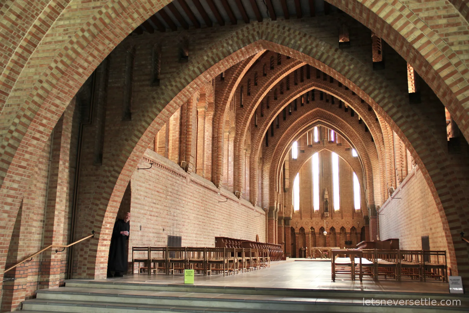 The brick nave of Quarr Abbey church with layered pointed arches and rows of wooden chairs, a robed monk standing near the entrance