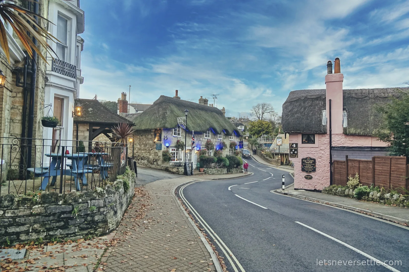 The curving road through Shanklin Old Village with a thatched stone pub hung with blue bunting on the left, the pink thatched teashop on the right and a bright blue sky above