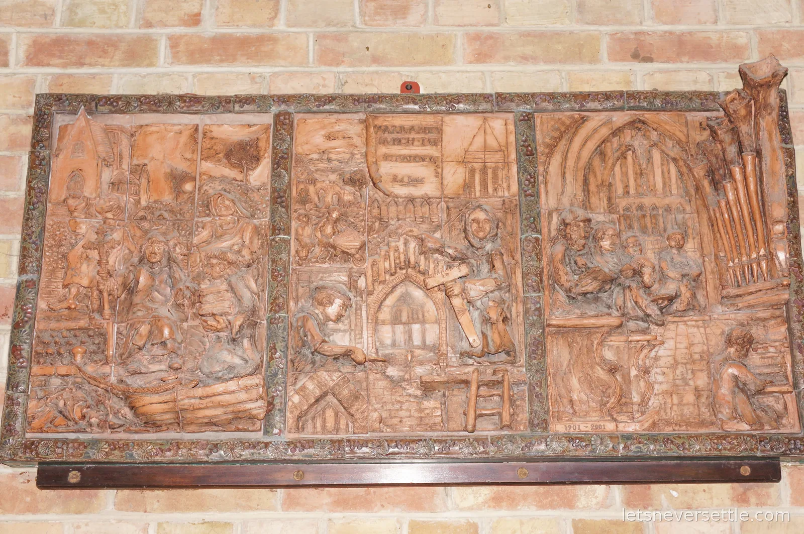 Terracotta relief panels on a brick wall showing monks building a church, with organ pipes and a congregation in the side panels