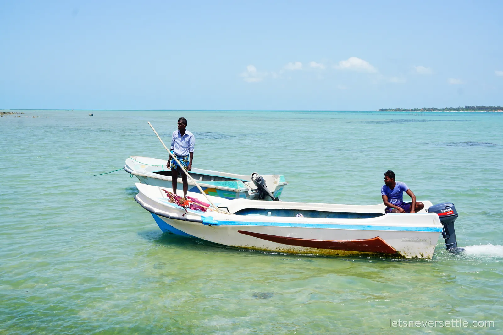 Two men standing in white fishing boats moored in clear turquoise shallow water under a bright sky