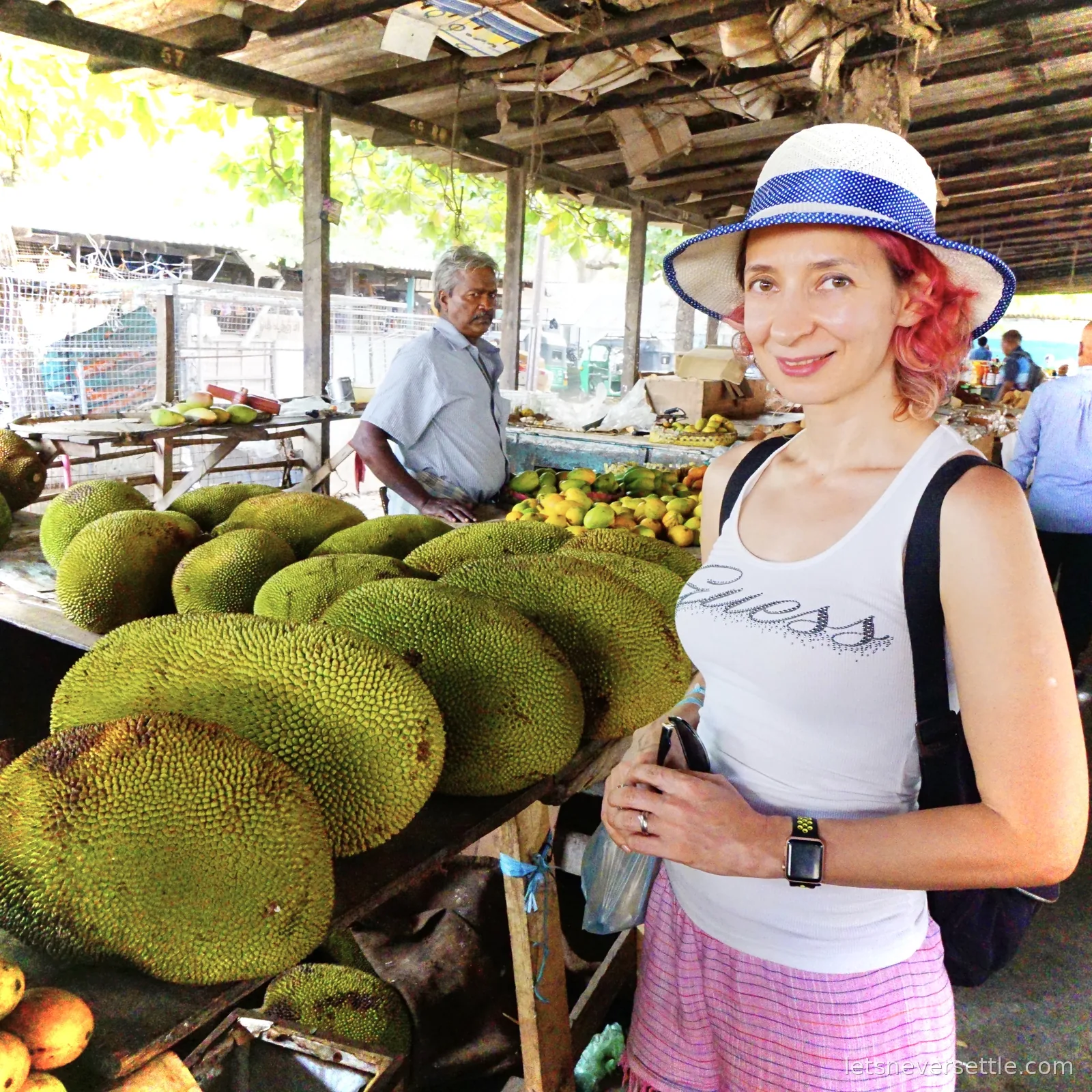Inga standing beside a market stall piled high with large green jackfruit, with a stallholder behind and mangoes on the next table