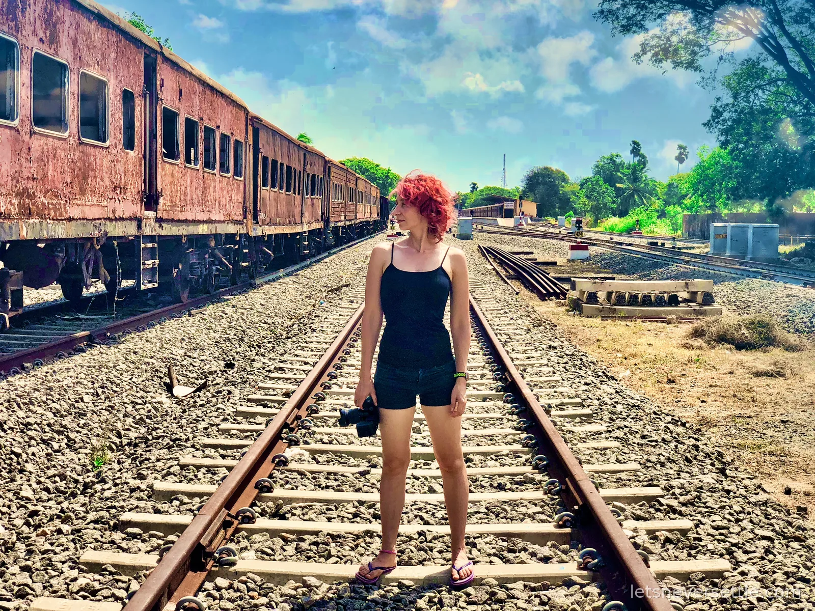 Inga standing on railway tracks beside a line of rusted, abandoned train carriages in a railway yard near Kankesanthurai
