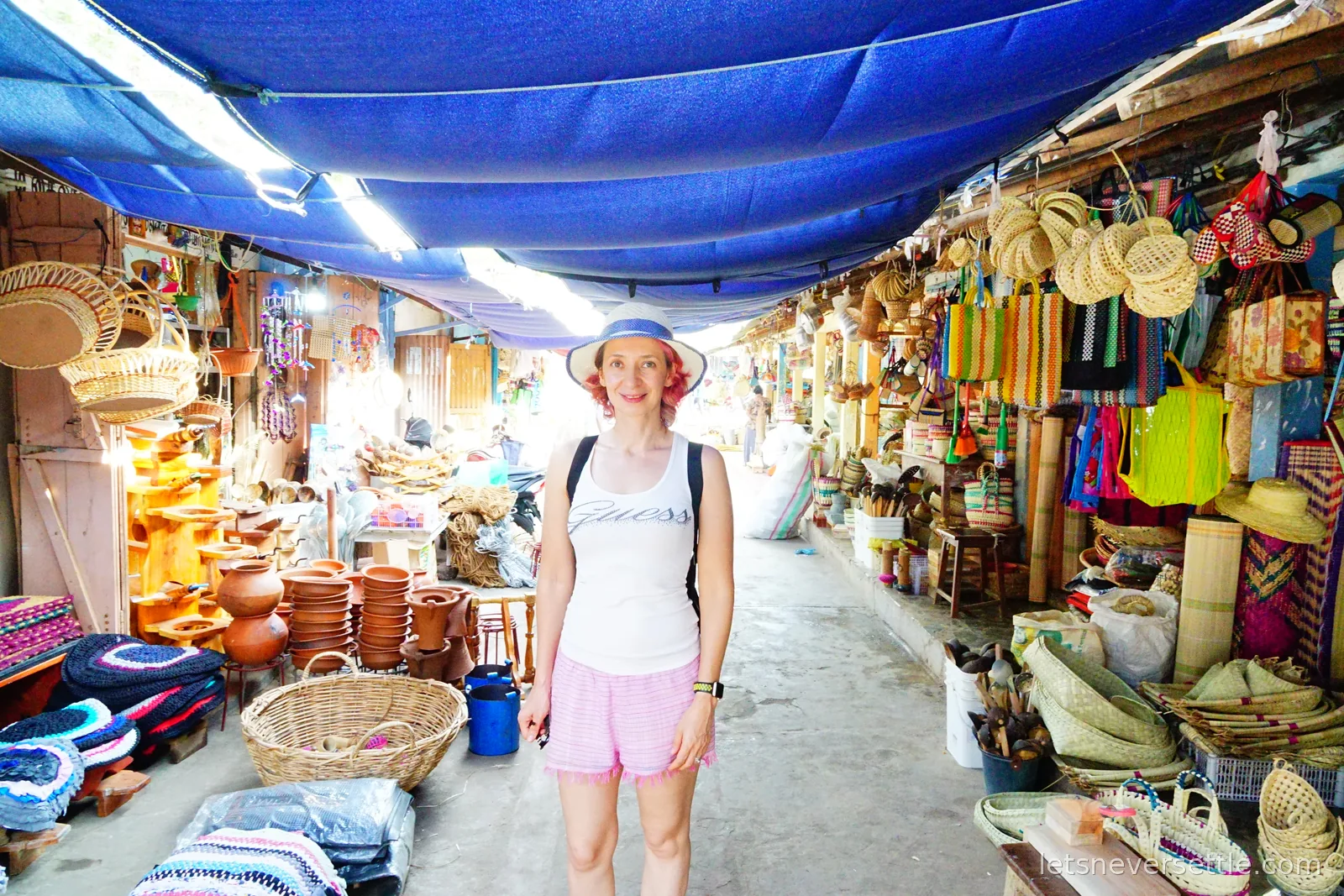 Inga in a sun hat standing in a market lane under a blue tarpaulin, lined with woven baskets, clay pots, mats and bags for sale