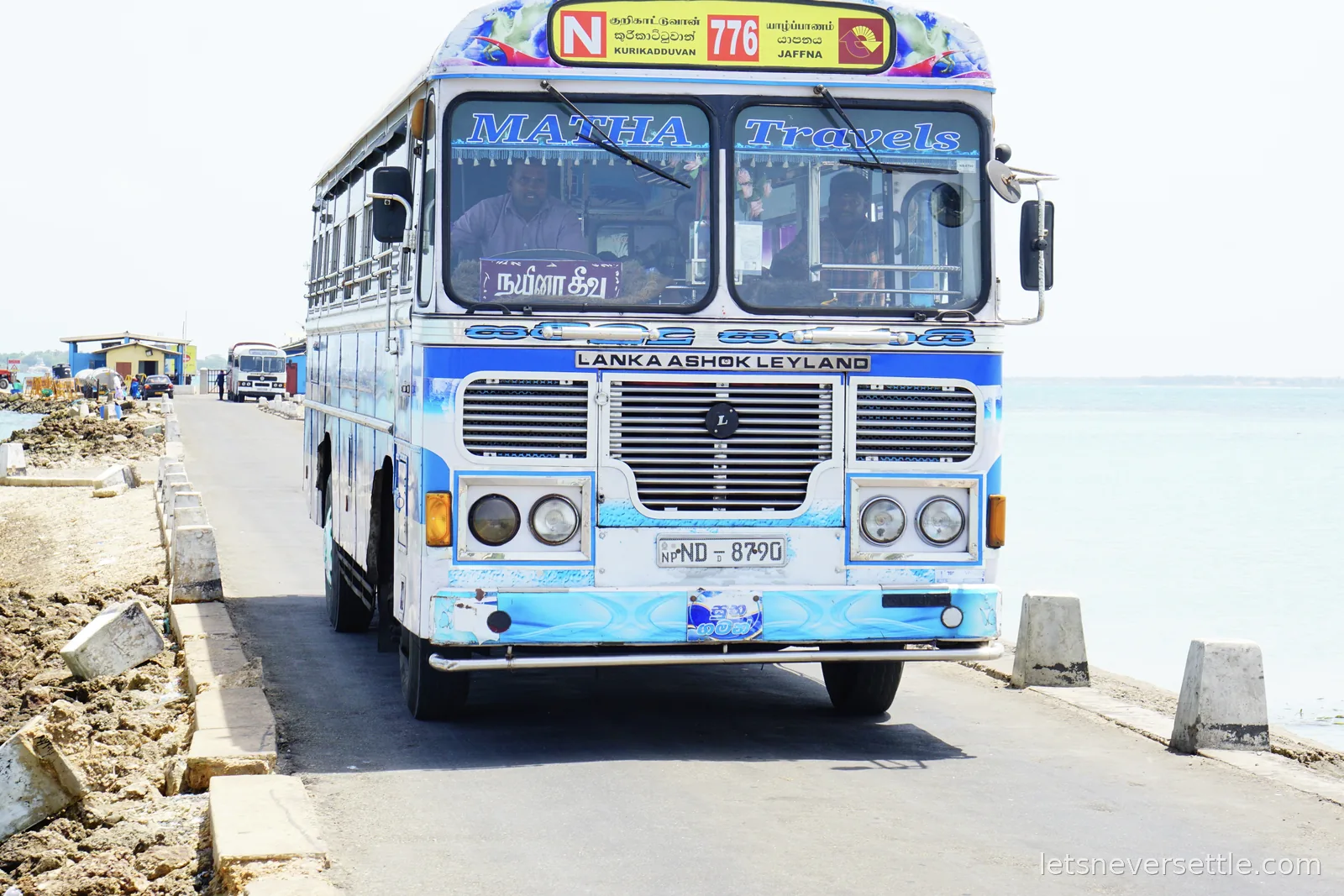 A blue and white Lanka Ashok Leyland bus lettered Matha Travels, route 776 to Jaffna, driving along a causeway road beside shallow sea