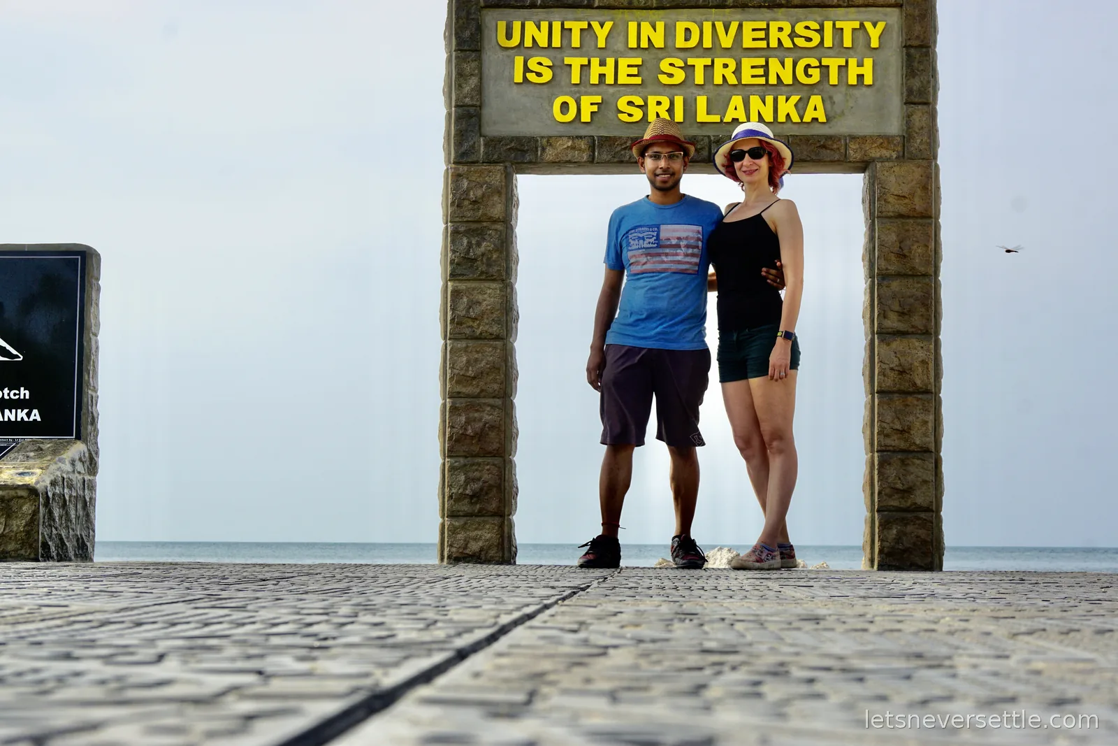 Inga and Dileep standing together under the tiled arch at Point Pedro, photographed from low down along the paved terrace with the sea behind them