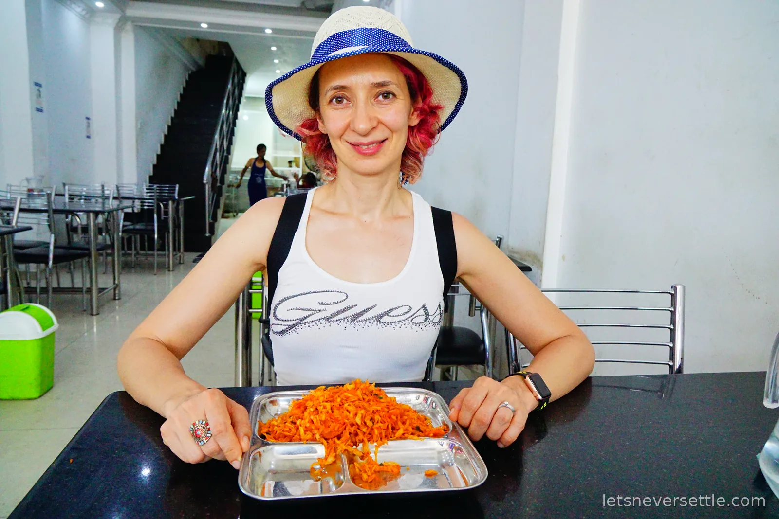Inga sitting at a table in a plain tiled cafe with a stainless steel tray of bright orange fried rice in front of her
