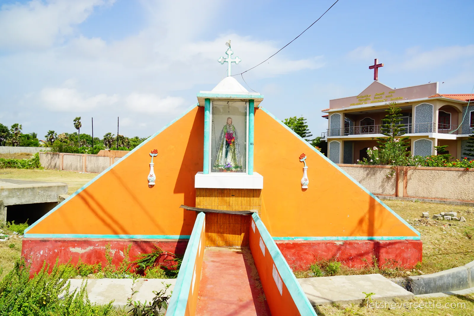 A small orange and red roadside shrine with a statue in a glass case at its centre, standing in front of a house topped with a cross