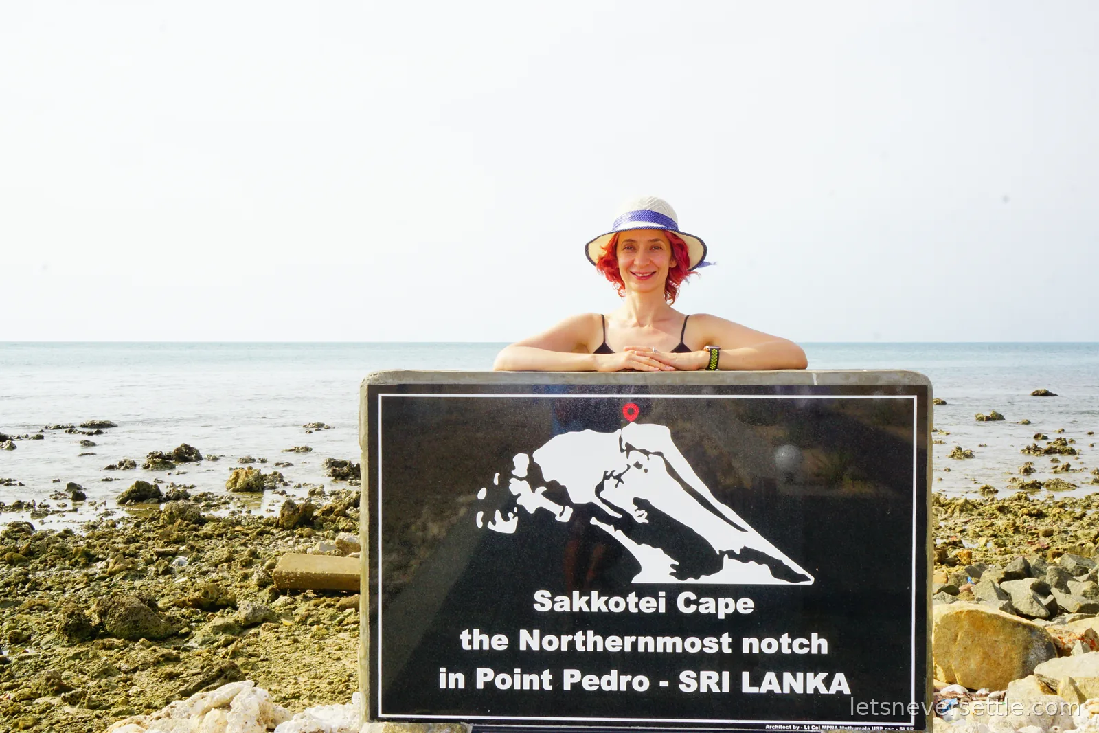 Inga in a sun hat leaning on a black granite plaque showing the outline of Sri Lanka with a red dot at the top, reading Sakkotei Cape the Northernmost notch in Point Pedro, with the sea behind