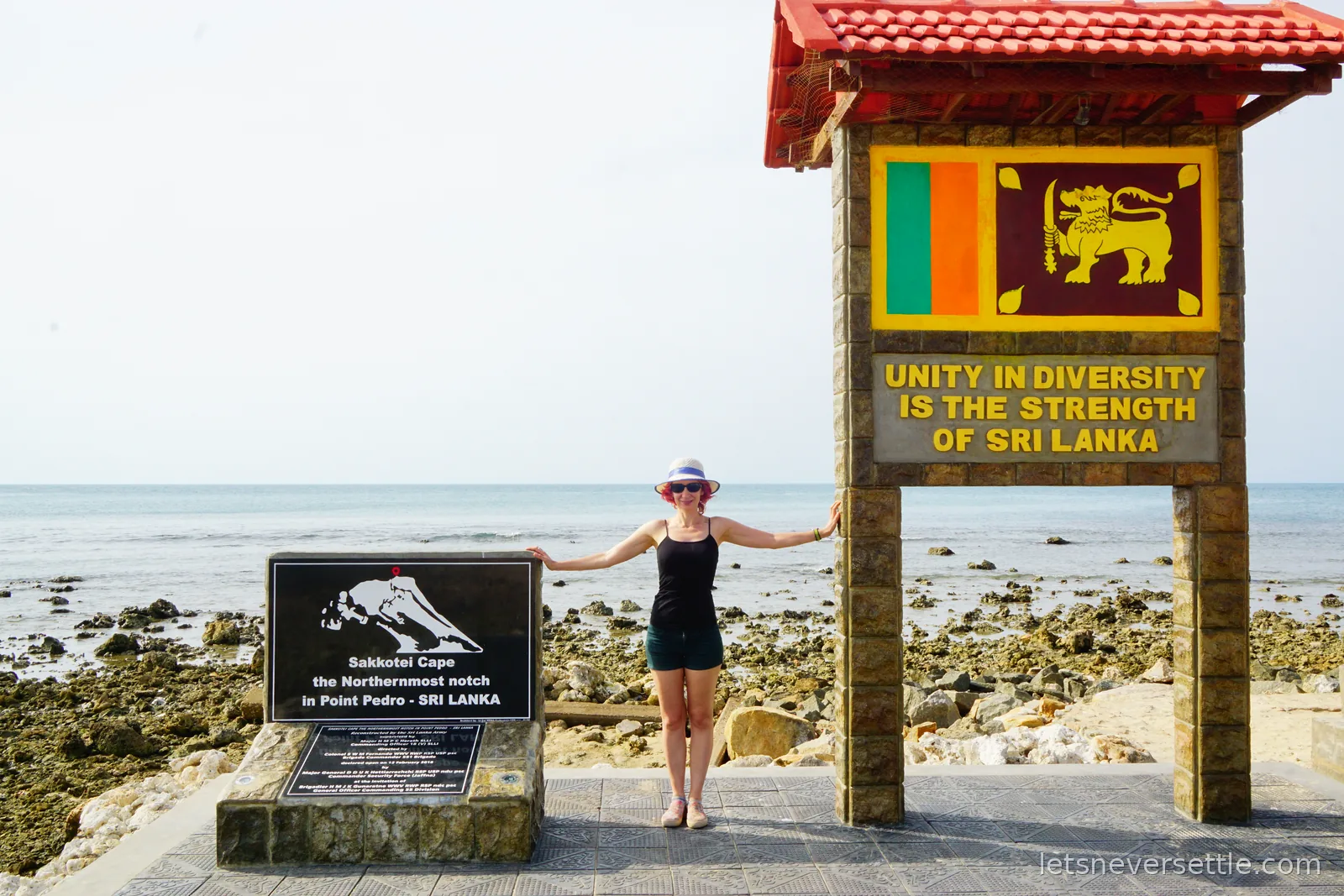 Inga standing with arms outstretched under a tiled arch at Point Pedro carrying the Sri Lankan flag and the words Unity in diversity is the strength of Sri Lanka, with the sea and coral rocks behind