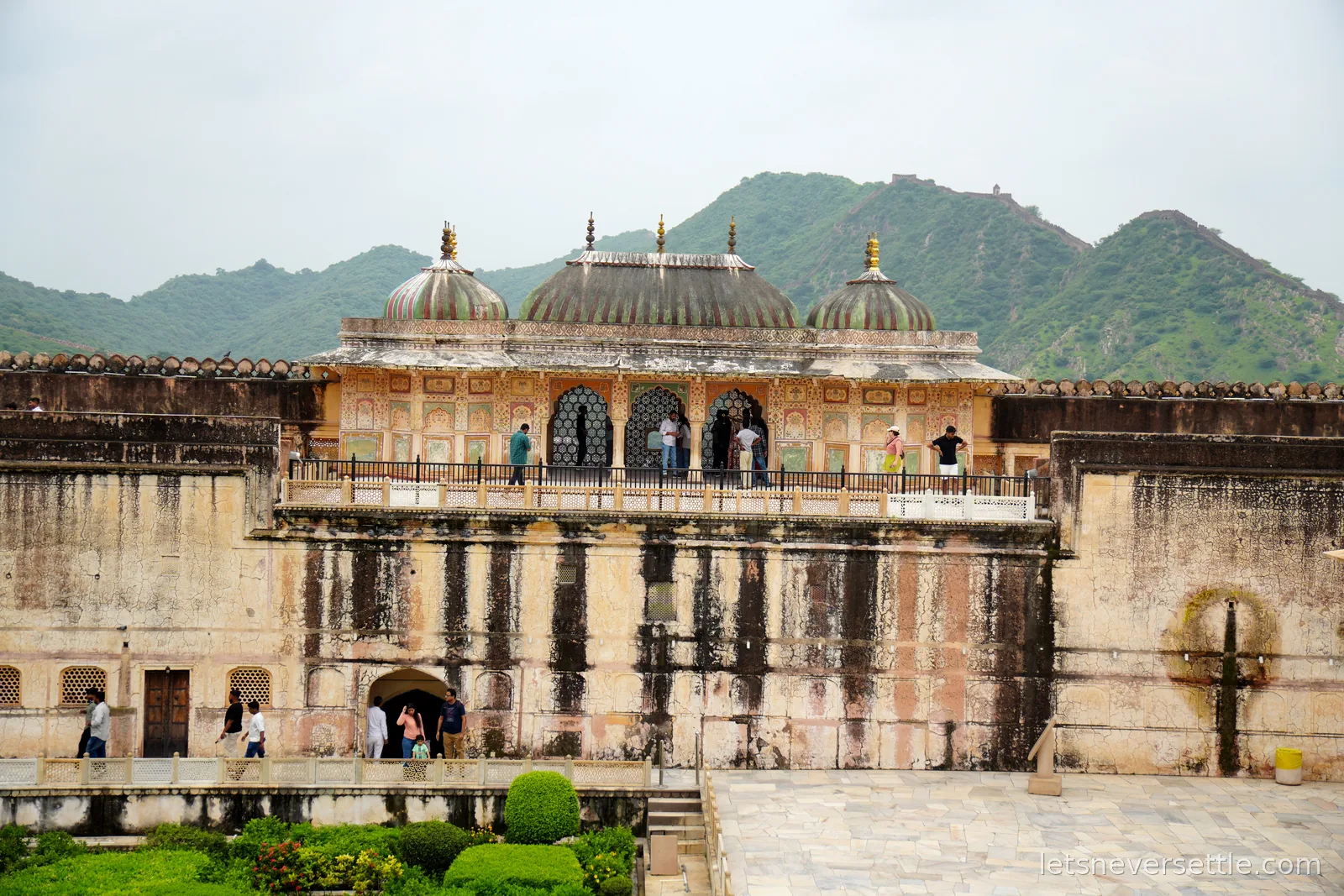 Domed pavilions above a weathered courtyard wall at Amber Fort, monsoon-green hills and old ramparts on the ridgeline behind