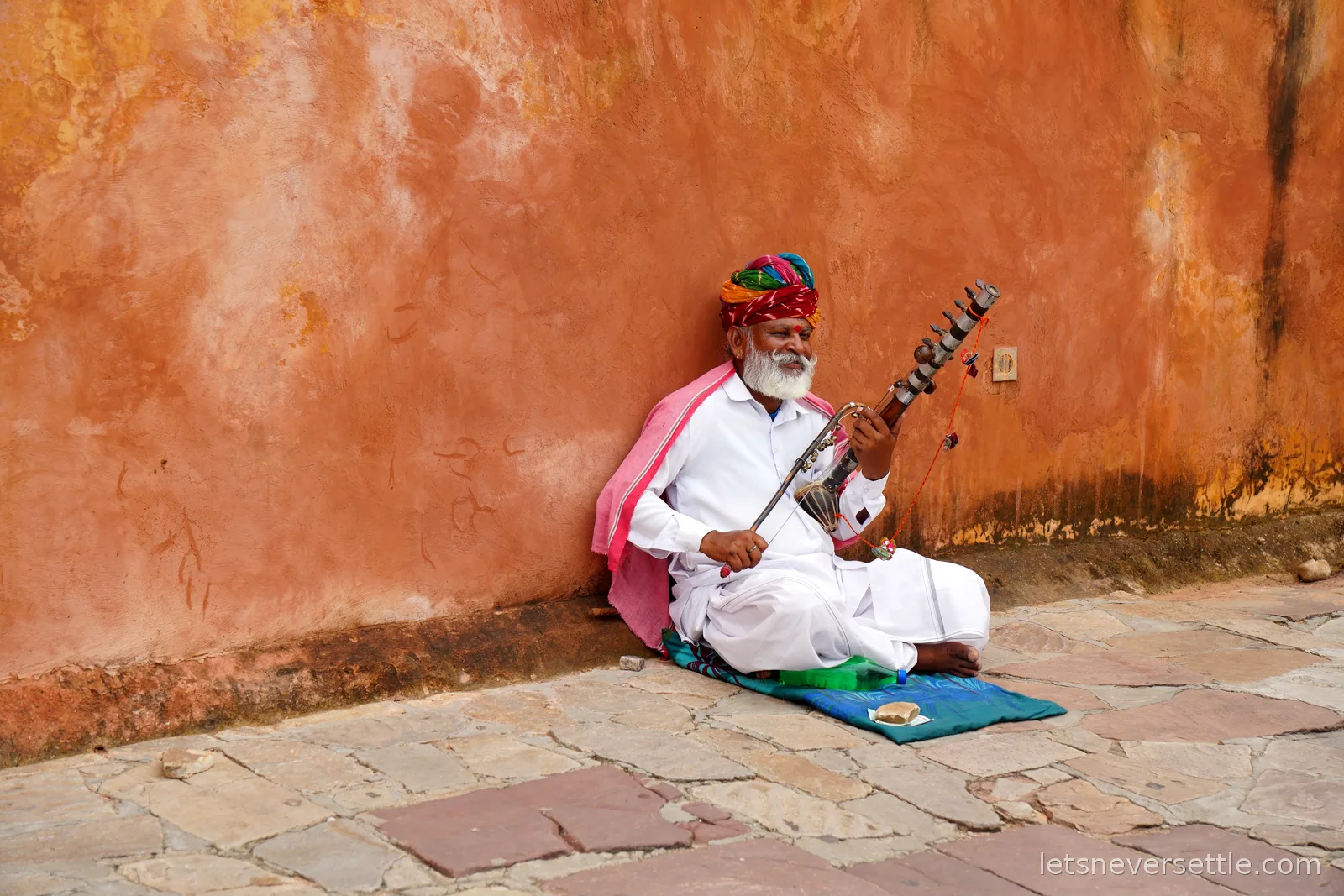 A musician in white with a bright turban playing a bowed string instrument while seated against an orange wall at Amber Fort