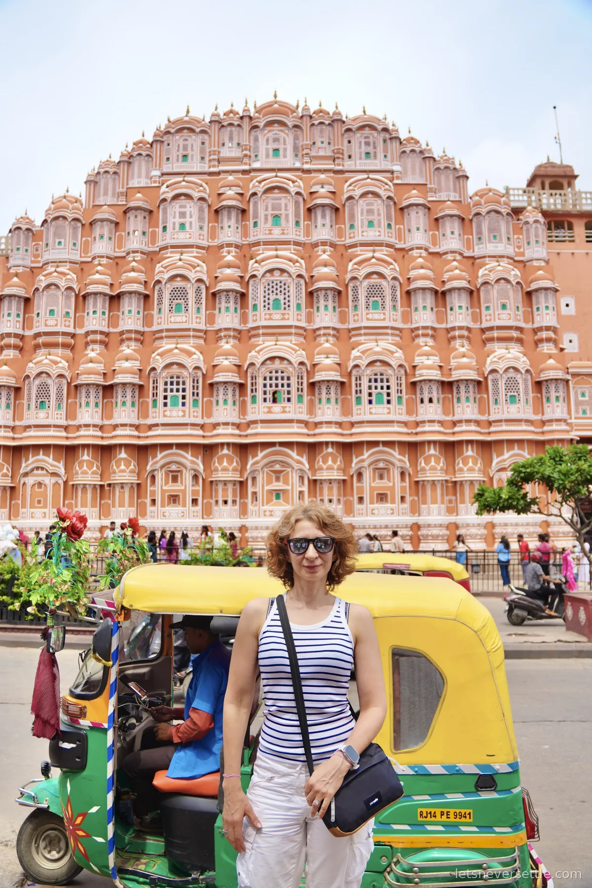 Inga in sunglasses in front of the pink honeycomb facade of Hawa Mahal in Jaipur, a green and yellow auto rickshaw at the kerb behind her