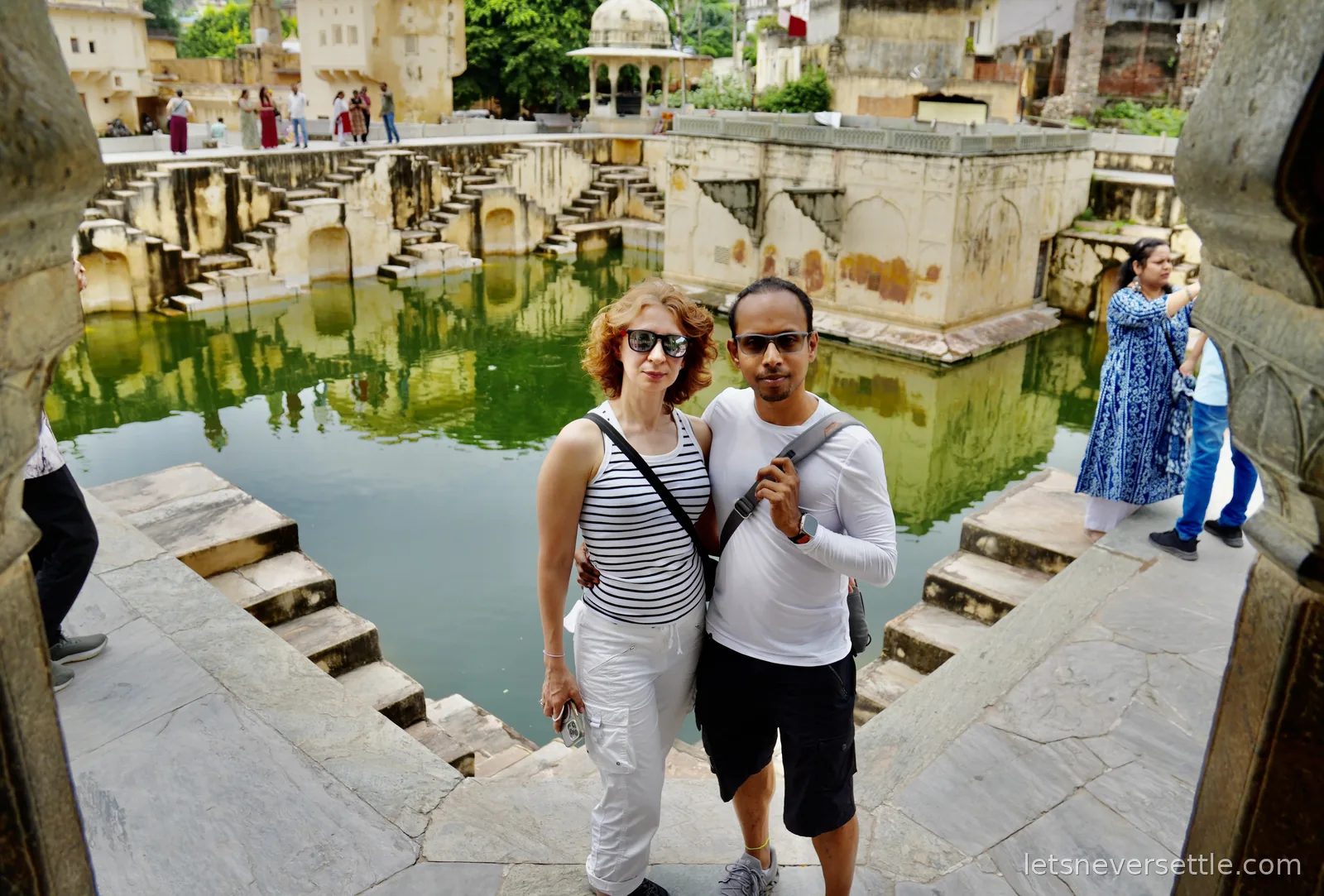 Inga and Dileep standing on the stone steps of Panna Meena ka Kund stepwell in Amer, zigzag staircases and green water behind them