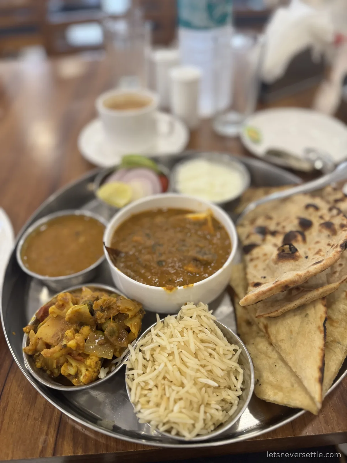 A steel thali tray with bowls of curry, dal, cauliflower sabzi, rice, naan, salad and a cup of chai behind it