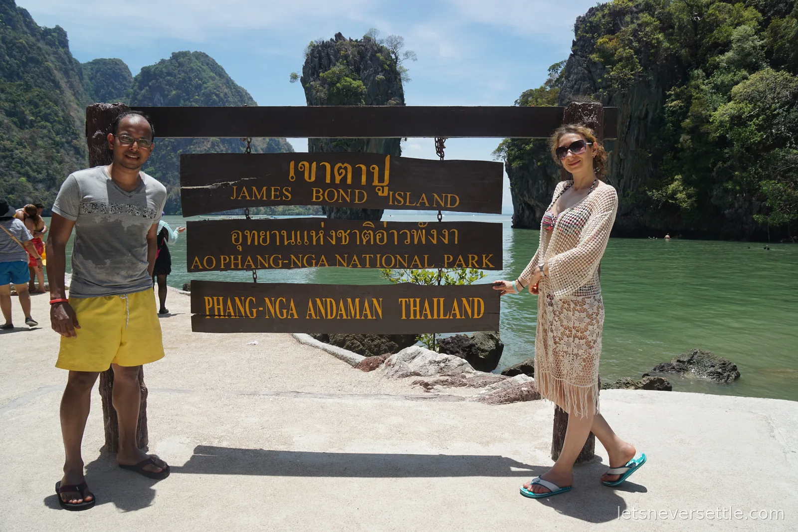 Inga and Dileep standing either side of a wooden signboard reading James Bond Island, Ao Phang-Nga National Park, Phang-Nga Andaman Thailand, with limestone cliffs and green water behind them