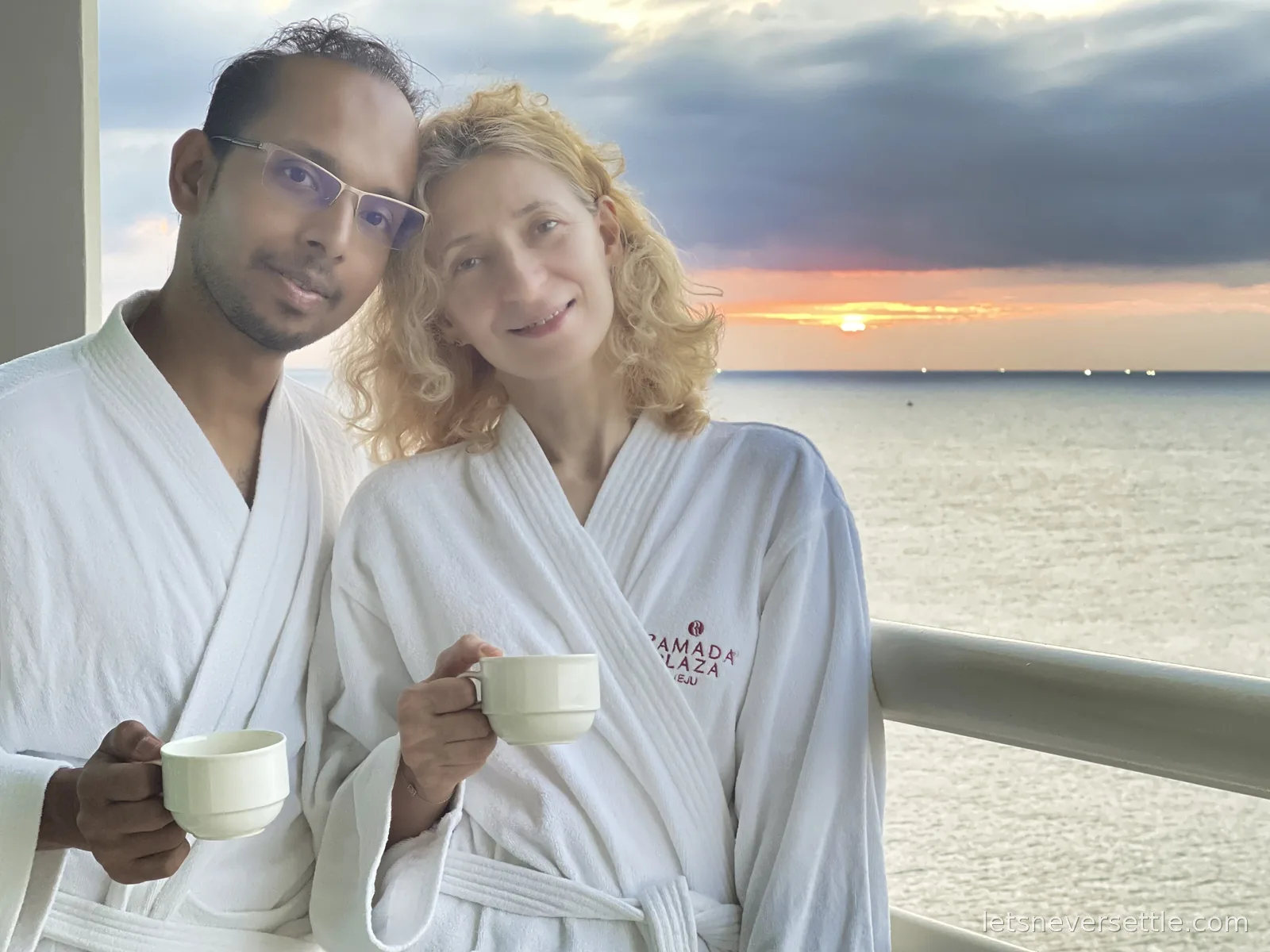 Inga and Dileep in white Ramada Plaza bathrobes holding coffee cups on a balcony as the sun sets over the sea
