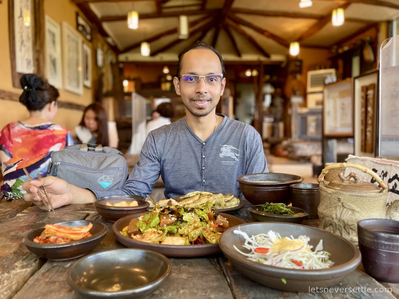 Dileep at a wooden table in a traditional Korean restaurant, earthenware pots, savoury pancakes and banchan in front of him