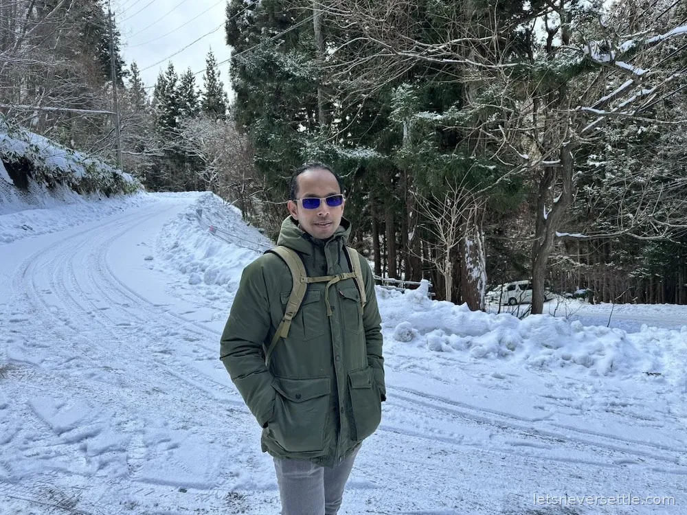Dileep in a green parka and sunglasses standing on a snow-covered road in Nagano with pine forest on both sides