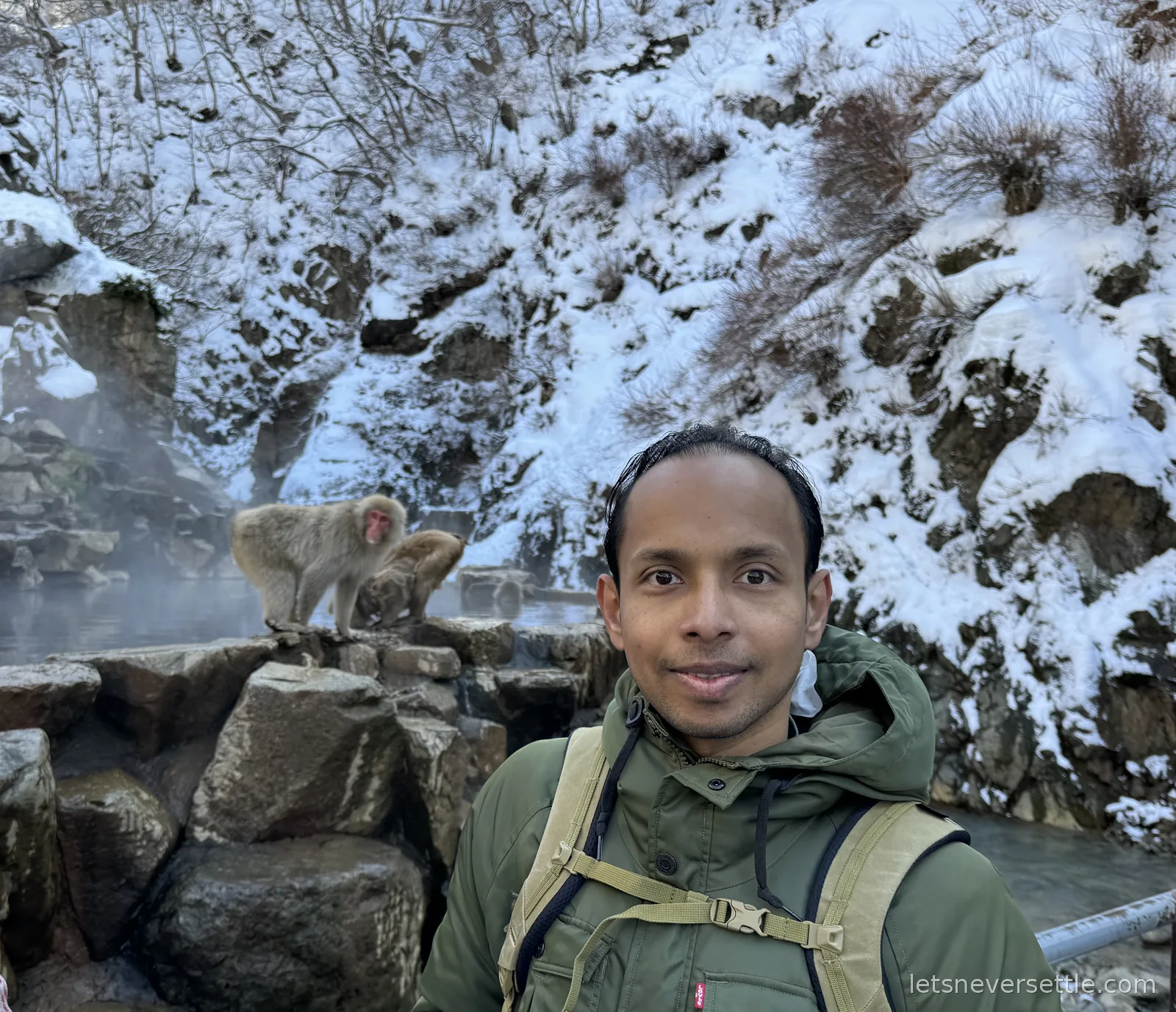 Dileep taking a selfie in front of snow-covered cliffs while two macaques groom each other on the rocks behind him